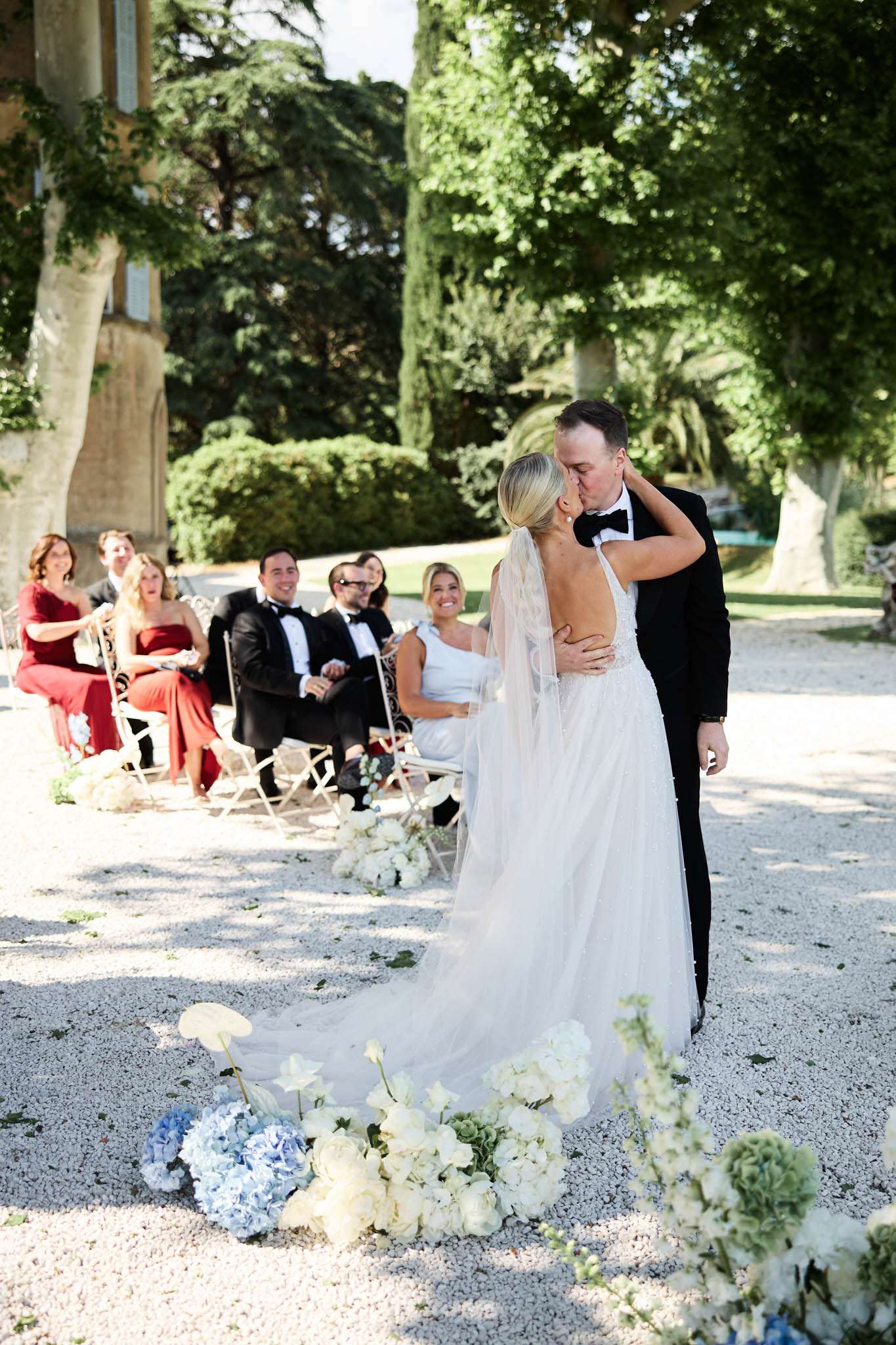 The bride and groom share their first kiss during an outdoor ceremony on a gravel allée, with approximately eight seated guests visible in the background reacting with smiles and laughter. The ceremony takes place in the grounds of a French château, with cypress trees and the stone façade of the building visible behind the guests. The bride wears a backless white tulle gown with a long cathedral train and a sheer veil, while the groom wears a black tuxedo with a black bow tie. The ceremony aisle is lined with clusters of white ranunculus, white hydrangeas, pale blue hydrangeas, and green hydrangeas arranged directly on the gravel, creating a loose floral border at ground level. Guests are dressed formally, with notable red and coral floor-length dresses among the women. The overall styling is classic and refined, with a white and blue floral palette. The shot is taken from behind the couple at a medium distance, capturing the full length of the bridal train and the floral aisle decor in the foreground.