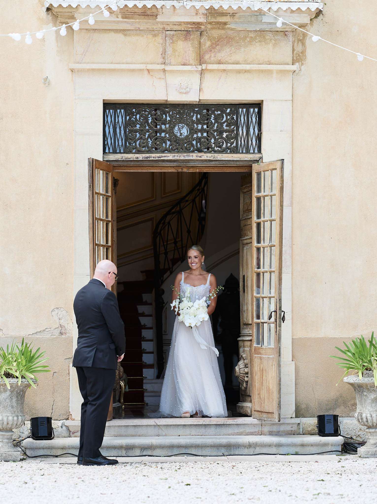 Bride in tulle gown with orchid bouquet appearing in chateau doorway as father stands with back to camera
