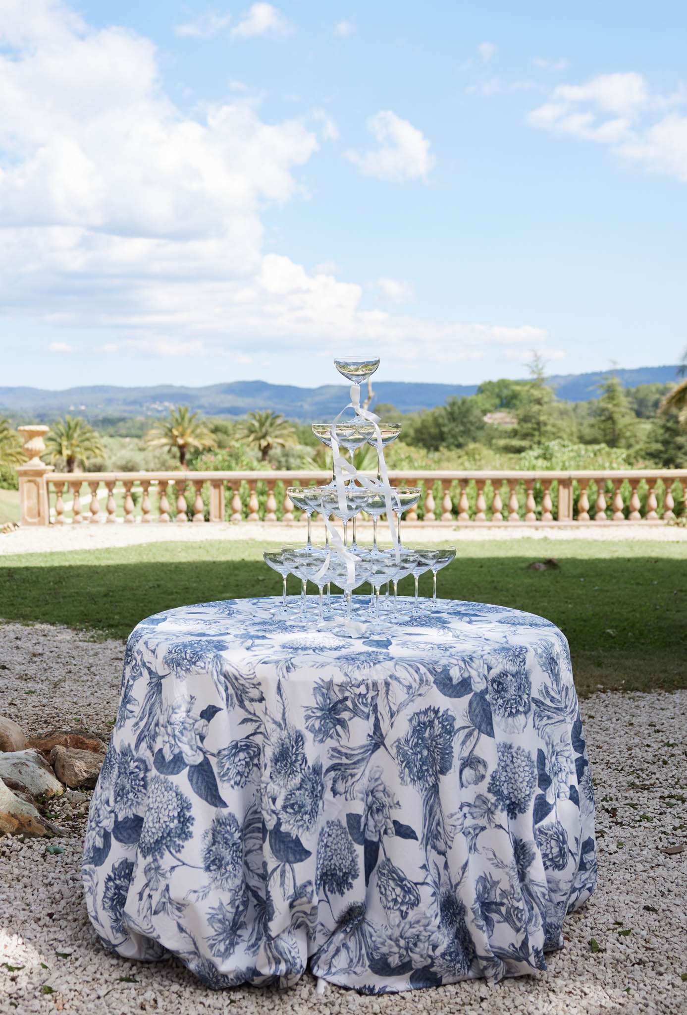 A champagne tower made up of coupe glasses arranged in a pyramid formation sits on a round cocktail table covered in a white and navy blue botanical toile-print tablecloth. A white ribbon winds decoratively up through the tower. The table is positioned on a gravel terrace outdoors, with a stone balustrade visible behind it overlooking a landscape of trees and rolling hills. The decor palette of navy and white gives the setup a classic French country aesthetic. Wide shot with no people present.