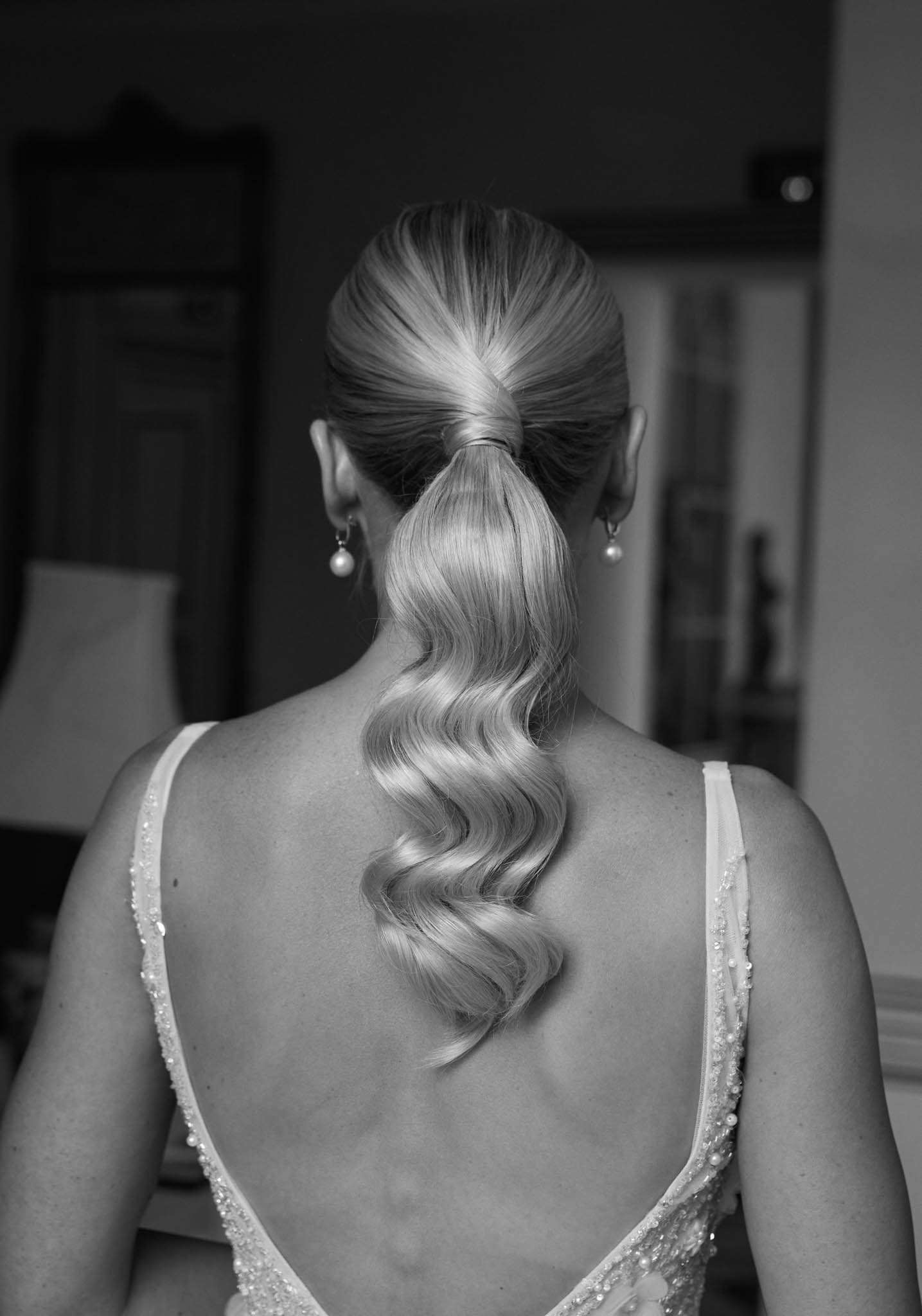 Black and white close-up of bride from behind showing low ponytail with barrel waves and pearl earrings