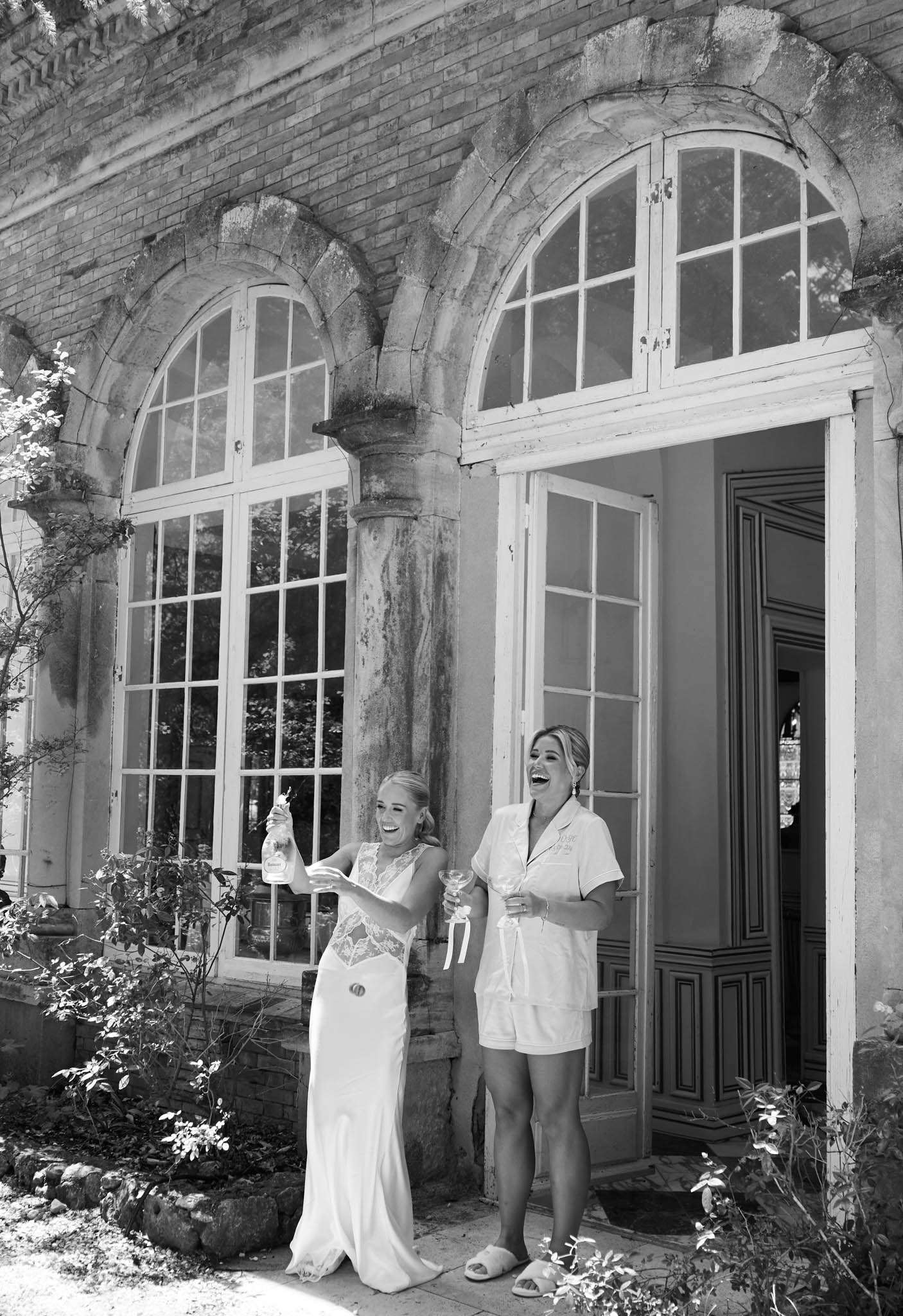 Black-and-white photo of bride and bridesmaid laughing while pouring champagne outside chateau facade