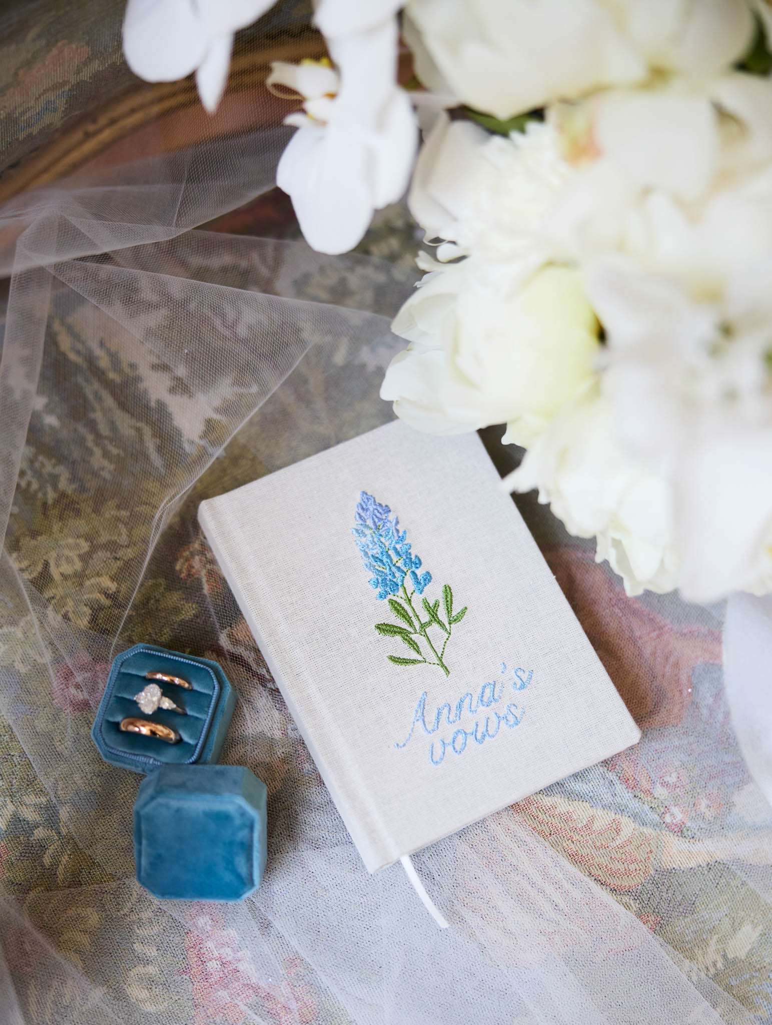 Linen vow book with blue embroidery, dusty blue velvet ring box with oval diamond, and white peony bouquet