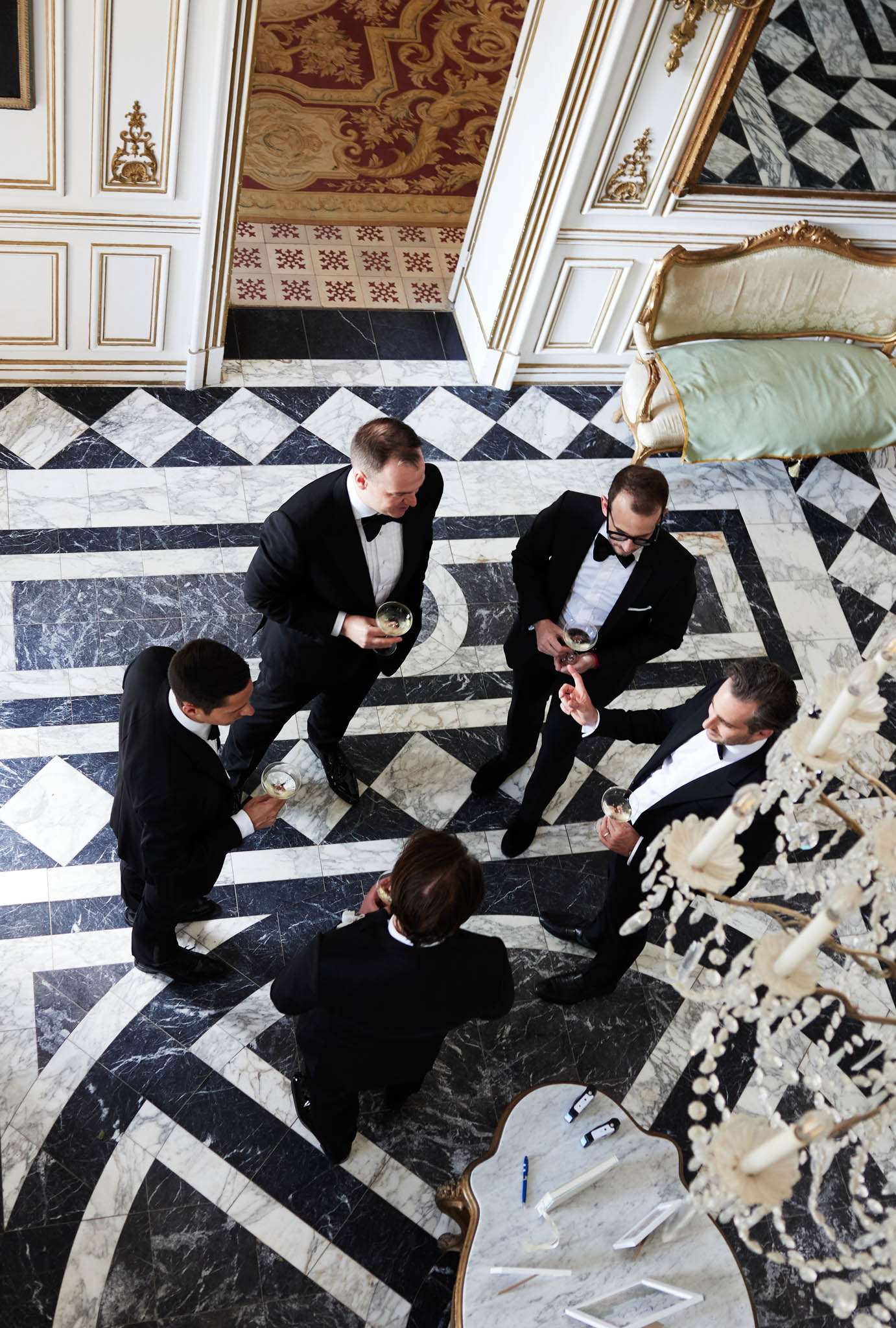 An overhead aerial shot captures five men in black tuxedos with black bow ties socializing during what appears to be a cocktail hour or pre-ceremony gathering inside a grand French château or palace-style venue. The men are holding champagne coupes and glasses, engaged in conversation in a loose cluster. The interior features a dramatic black and white marble geometric floor with diamond and square patterns, white paneled walls with gold gilt moldings, a glimpse of an ornate red and gold ceiling fresco, and a mint green upholstered gilt-framed settee visible in the upper right corner. A crystal candelabra with white taper candles and an ornate gold-legged side table are partially visible in the lower right. The classic black-tie dress code and formal interior decor reflect a high-end, formal wedding aesthetic. Potential venue feature image.