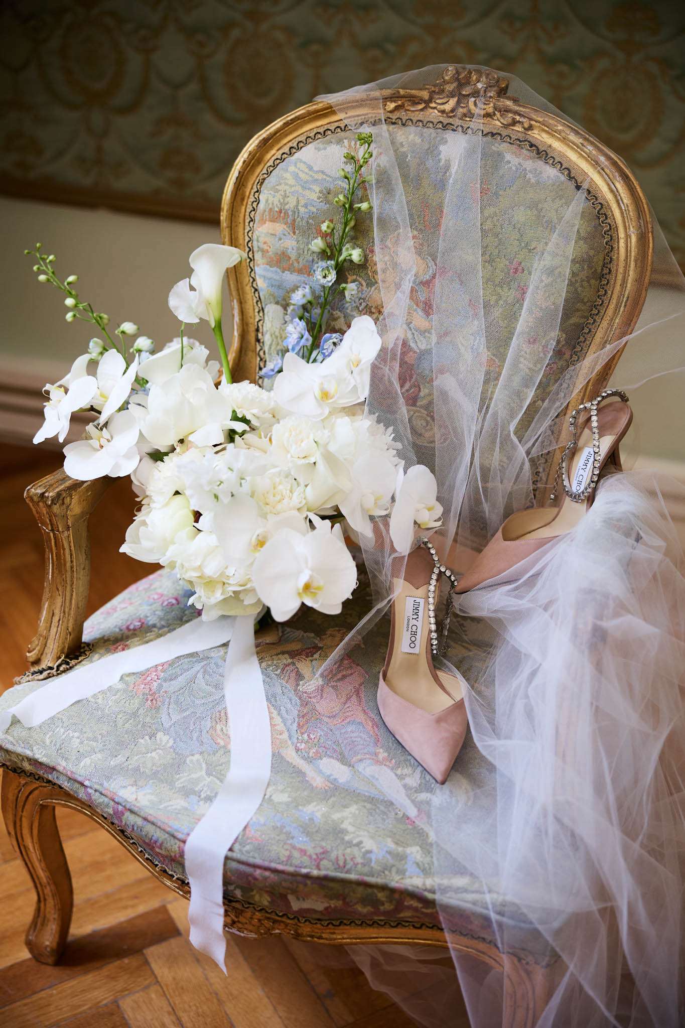Bridal flat lay on antique armchair with white peony bouquet, blush pink heels, and tulle veil