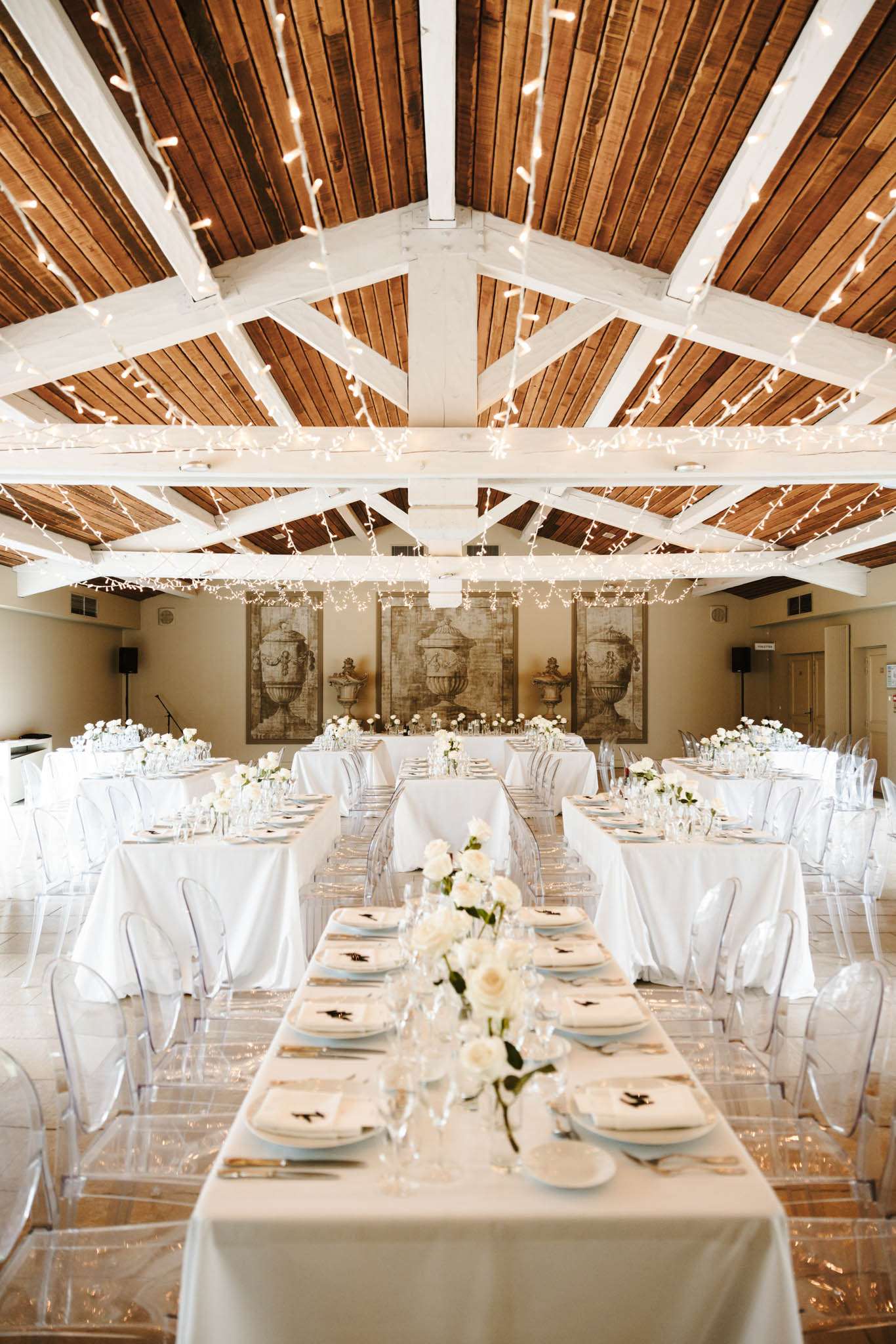 Barn reception with fairy-lit vaulted beams ghost chairs cream rose centerpieces and sepia wall panels