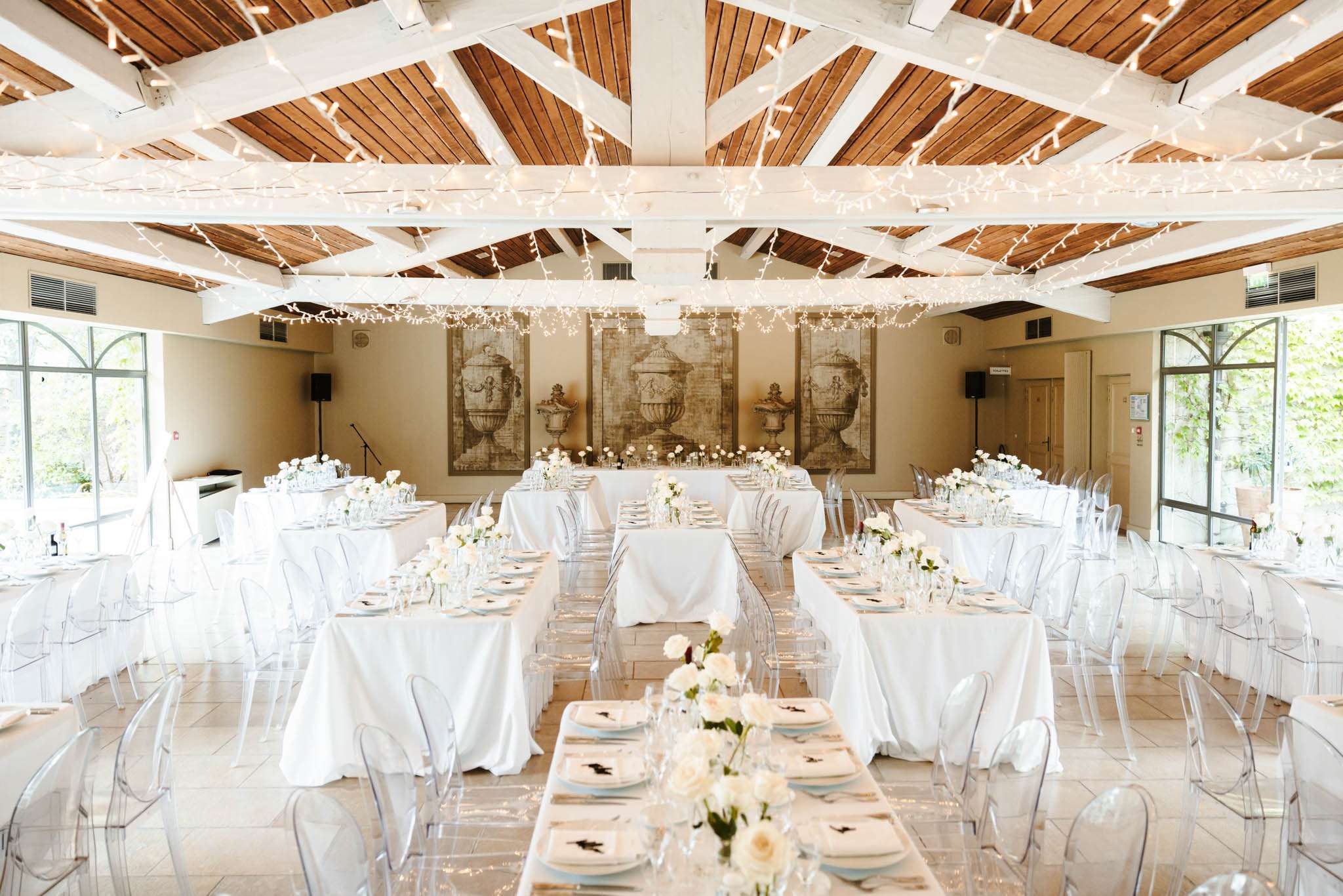 A wedding reception room set up for a seated dinner, photographed from a wide-angle perspective showing the full layout of the space. The indoor venue features a vaulted ceiling with white-painted wooden beams and warm wood paneling, draped with dense fairy lights that run the length of the room. Tables are arranged in long rectangular rows covered with white linen tablecloths, set with clear glassware, light blue charger plates, and white dinner plates with a dark botanical print motif. Low centerpieces of cream and ivory roses are placed at intervals along each table. All seating consists of clear acrylic ghost chairs. At the far end of the room, a head table is positioned in front of a large triptych artwork depicting classical urns in muted sepia tones, flanked by arched floor-to-ceiling windows on both sides that let in natural daylight. The overall decor palette is white, cream, and clear with pale blue accents, giving the space a clean, modern-classic aesthetic. Potential venue feature image.