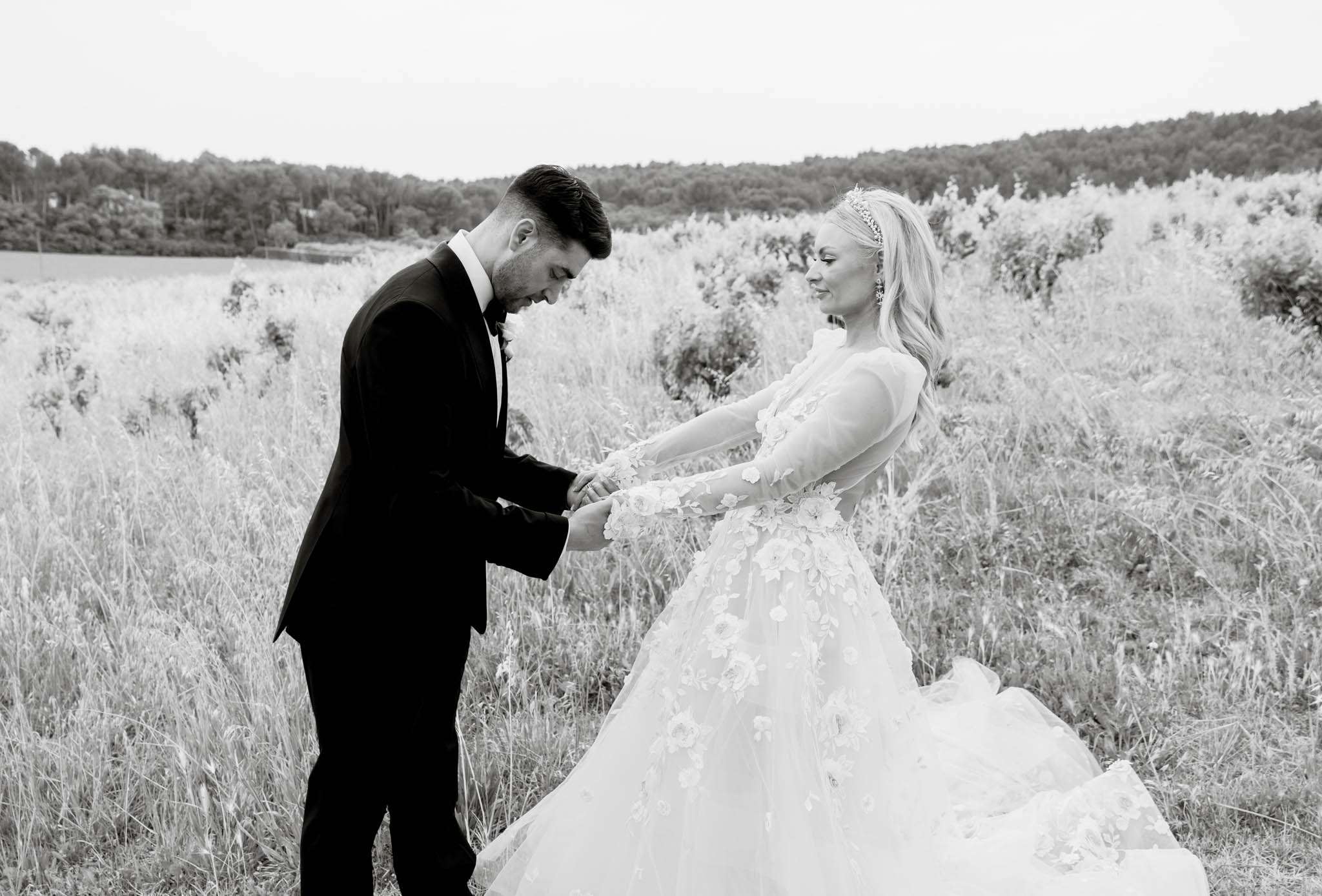 This is a black-and-white couple portrait taken outdoors in a countryside setting, likely among vineyard rows or tall wild vegetation. The bride and groom are holding hands and facing each other; the groom, in a dark tuxedo with bow tie, looks down at their joined hands while the bride looks toward him with a slight smile. The bride wears a full ballgown with sheer long sleeves and heavily appliquéd three-dimensional floral detailing across the bodice and skirt, along with a delicate tiara and drop earrings, with long wavy hair. The image is rendered in high-contrast black and white, with the deep tones of the groom's suit contrasting sharply against the bright whites of the bride's gown and the light-toned vegetation behind them. The composition is a mid-wide portrait shot with the landscape extending into the background.