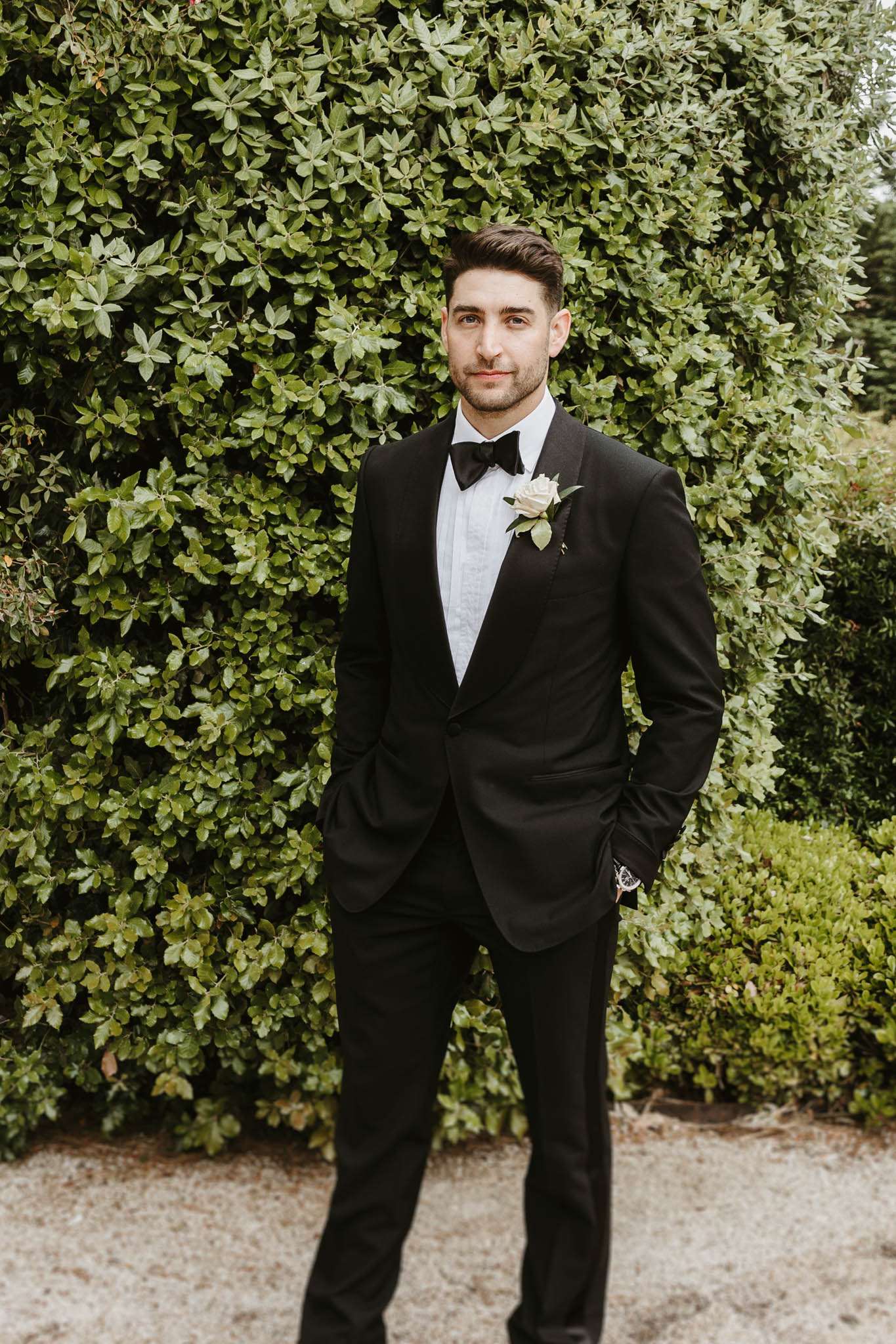 A solo portrait of the groom standing outdoors in front of a dense hedge backdrop, with gravel visible at ground level. He is wearing a black tuxedo with satin lapels, a white dress shirt, a black bow tie, and a white rose boutonniere with small green leaves. His hands are in his pockets and he is looking directly at the camera with a neutral expression. A watch is visible on his left wrist. The shot is a full-length portrait with a classic, formal styling aesthetic.