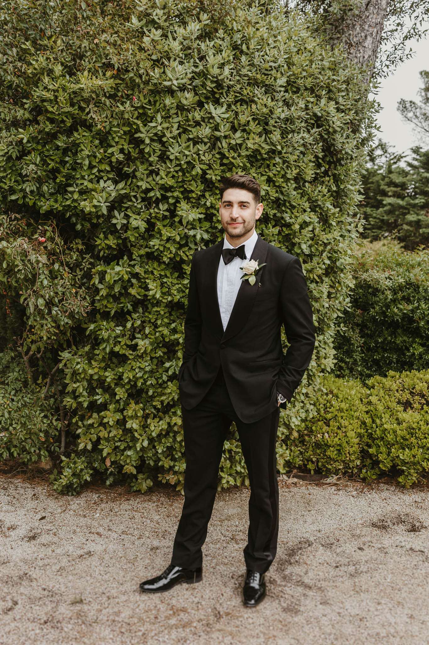 Groom in black tuxedo with bow tie and cream rose boutonniere standing with hands in pockets before green hedge