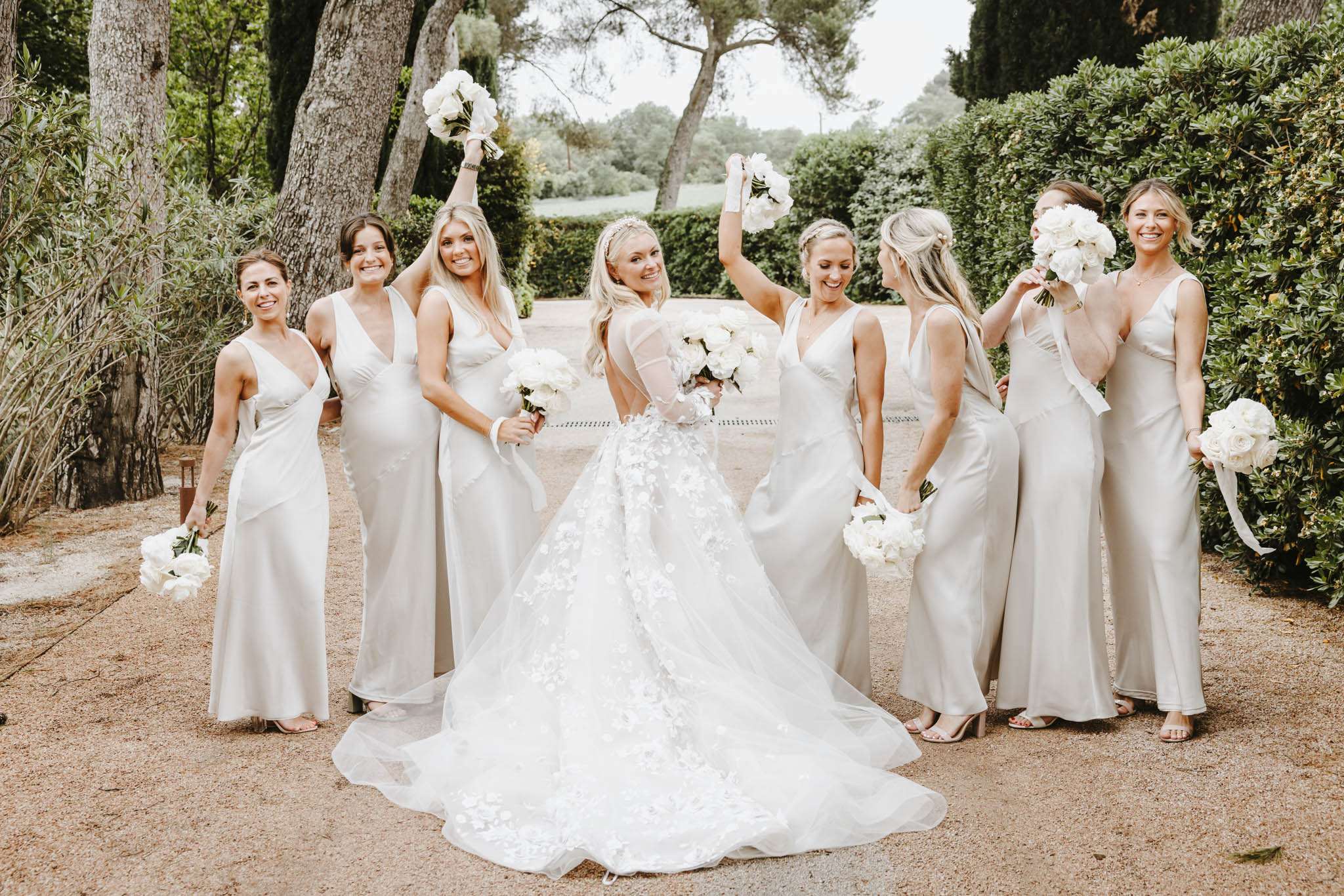 A bridal party portrait taken outdoors on a gravel path surrounded by manicured hedges and tall trees. The bride stands at the center facing away from the camera, showing the back of her white ball gown, which features a full tulle skirt with three-dimensional floral appliqué detailing and a long train; she wears long sheer sleeves and a pearl or crystal headband. She is surrounded by six bridesmaids wearing floor-length champagne-sage satin slip dresses with V-necklines, all holding white bouquets composed of large peonies or garden roses with trailing ribbons — several are raising their bouquets in celebration. The overall color palette is white and warm champagne-grey, consistent with a classic, clean aesthetic. The shot is a medium-wide group portrait with the bride's back as the focal point.