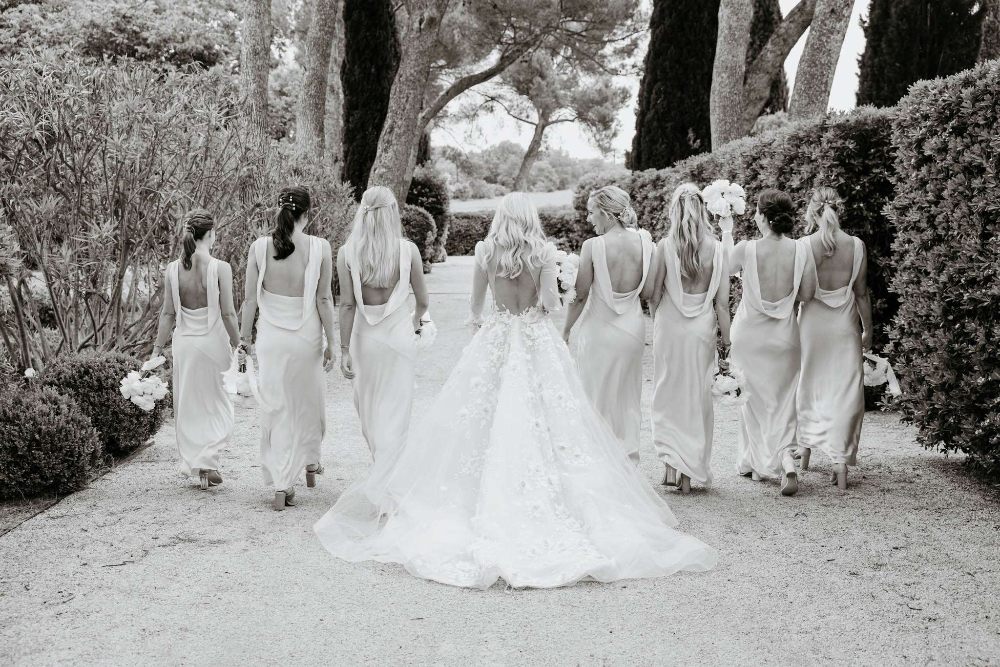 Black and white rear view of bride and seven bridesmaids walking along a formal garden path with hedges