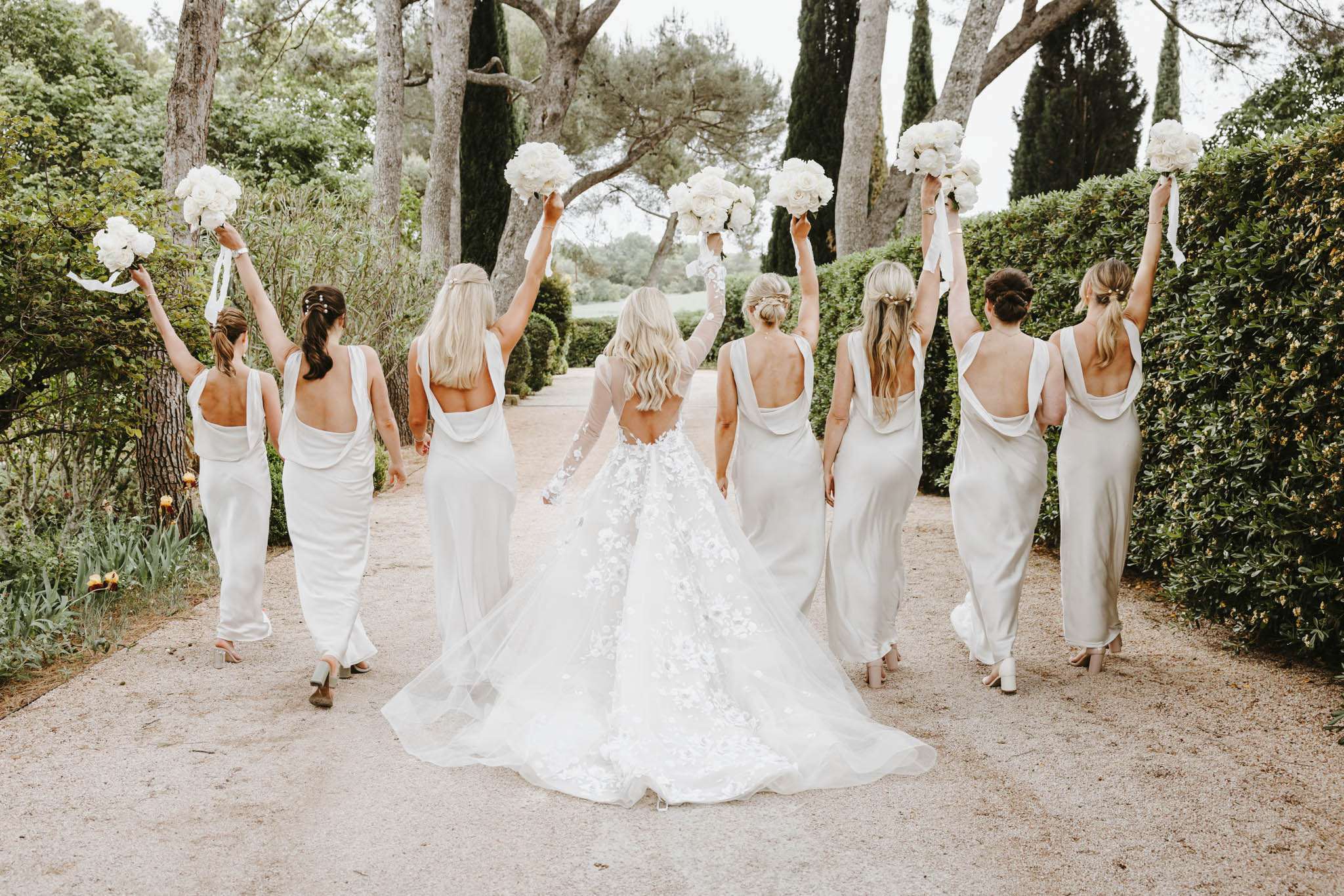A bridal party portrait taken from behind, showing the bride and seven bridesmaids walking away down a gravel path lined with trimmed hedges and tall trees. The bride wears a white ball gown with a deep open back, sheer long lace sleeves, and a full 3D floral appliqué skirt with a long train. The seven bridesmaids are dressed in matching ivory satin cowl-back slip dresses with a deep draped open back. All eight women have their bouquets raised overhead — the bouquets are composed of white peonies or garden roses with white ribbon streamers. The overall color palette is an all-white and ivory scheme with a classic, modern aesthetic. This is a wide-angle candid-style group portrait shot outdoors at a French estate or château garden.