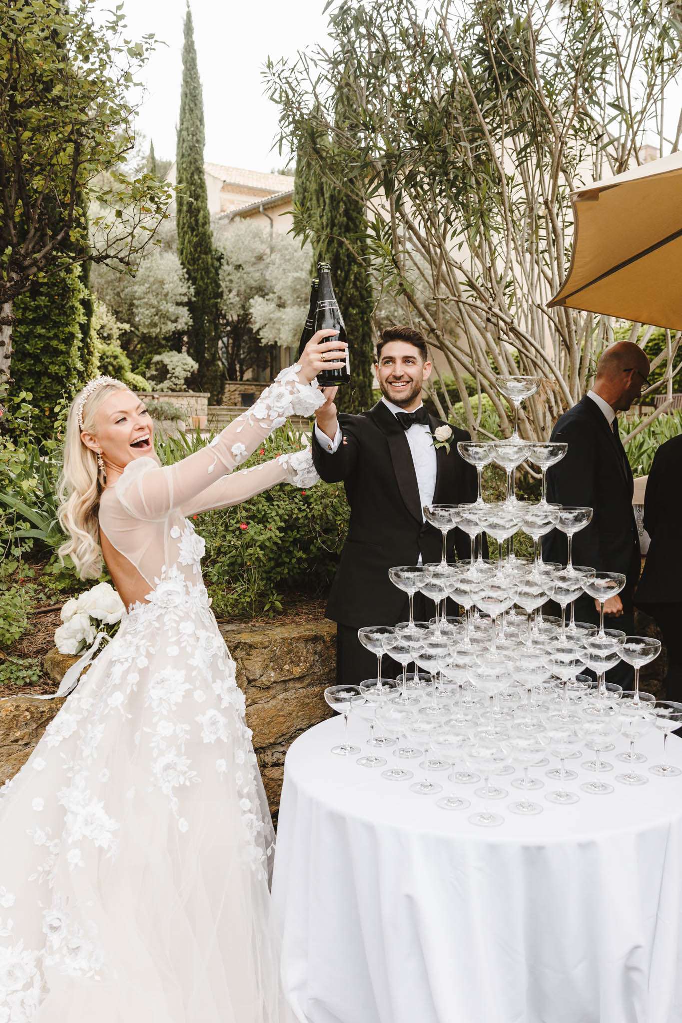 Bride in floral applique ballgown pours champagne into coupe tower as groom watches at Provencal venue