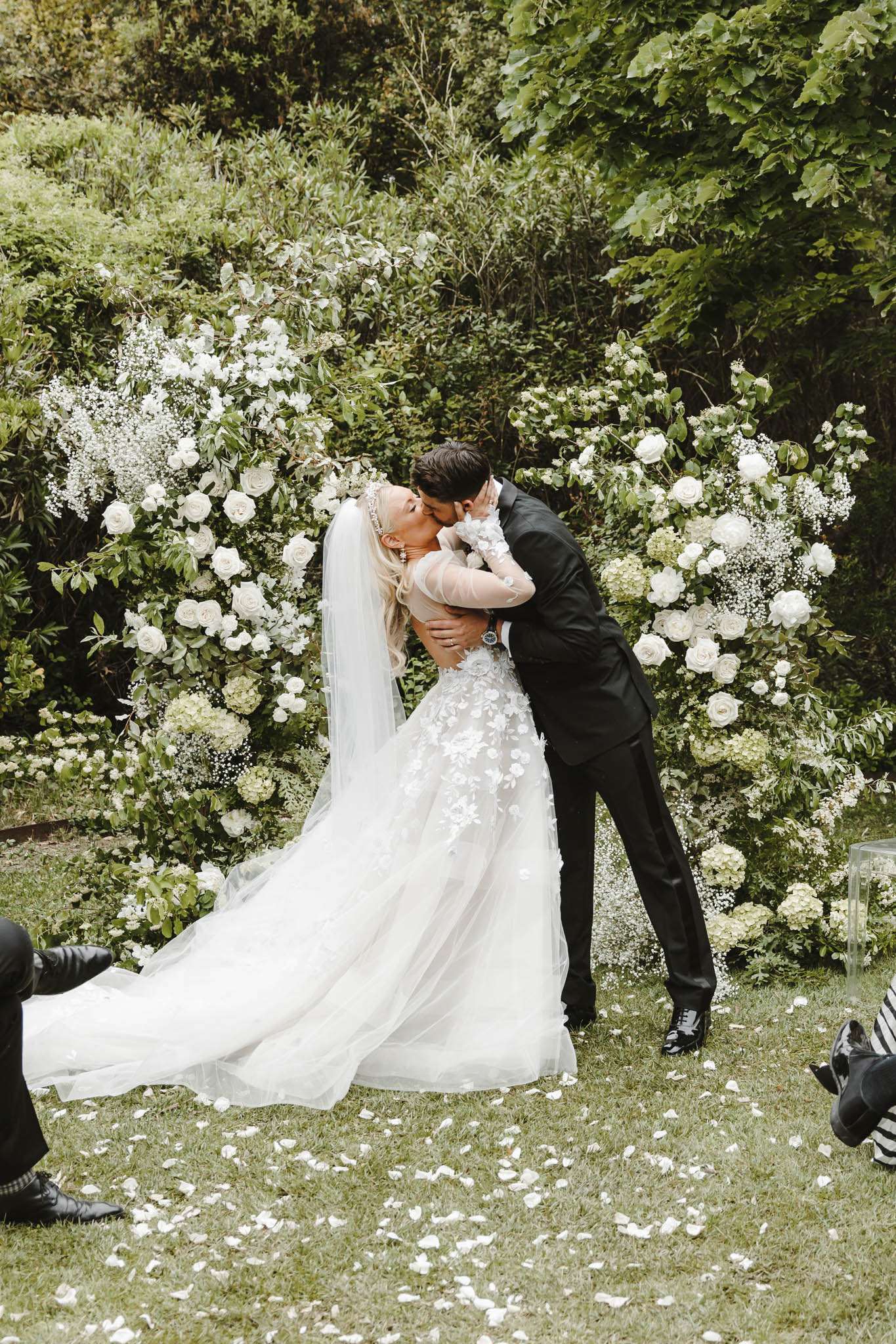 Couple's first kiss before white rose and hydrangea column arrangements with petals scattered on grass