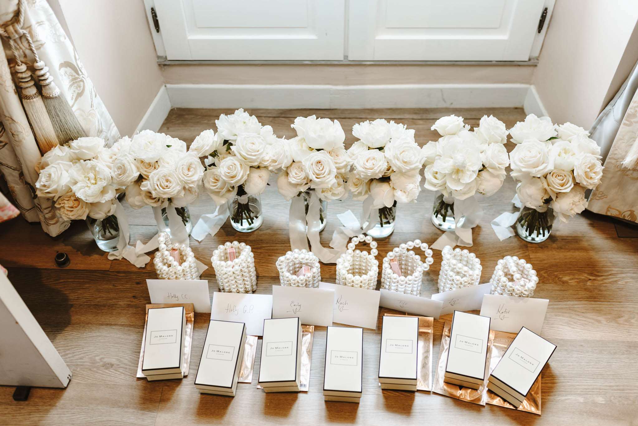 A flat lay-style getting-ready detail shot taken from above, showing seven bridesmaids' gift sets arranged in a row on a wooden floor in front of a window. Each set includes a bouquet of white roses and white peonies in a glass vase tied with a white satin ribbon, a pearl beaded cuff bracelet, a personalised name card, and a Jo Malone fragrance box in white and black packaging. The Jo Malone boxes are placed on rose gold gift bags. The bouquets are composed entirely of white and ivory roses and peonies, maintaining a consistent all-white floral palette across all seven arrangements. The pearl bracelets are stacked cuff-style with layered rows of white pearls. Patterned curtains with tassel tiebacks are visible on either side of the frame, suggesting an interior room setting. The overall gift aesthetic follows a classic, all-white and pearl colour palette.