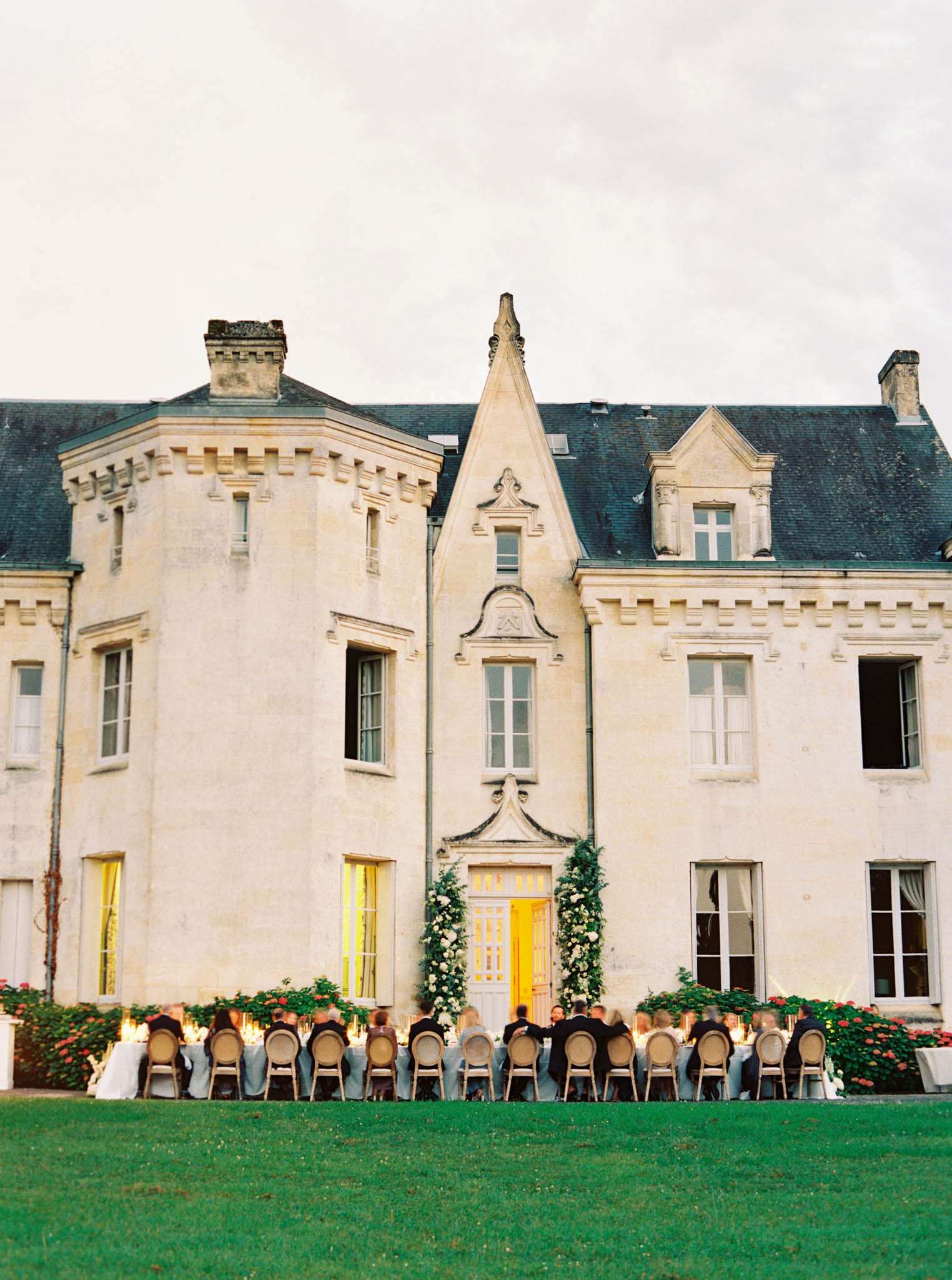 An outdoor wedding reception dinner is taking place directly in front of a French château, with approximately 30–40 guests seated at a long rectangular table arranged parallel to the building's façade. The château features cream/ivory stone architecture with a slate-grey mansard roof, decorative dormer windows, turret corners, and ornate carved detailing around the central entrance. The front door is framed by a lush floral installation of white blooms and greenery climbing the doorway, and warm golden light glows from inside the entrance and ground-floor windows, creating a lantern-like effect at dusk. The table is lined with candles and what appear to be tall floral centerpieces, with round-backed wooden dining chairs and white table linens visible. The classic, formal styling is consistent with a French château reception aesthetic. Wide shot taken from across the lawn, capturing the full building facade and the full length of the dining table. Potential venue feature image.