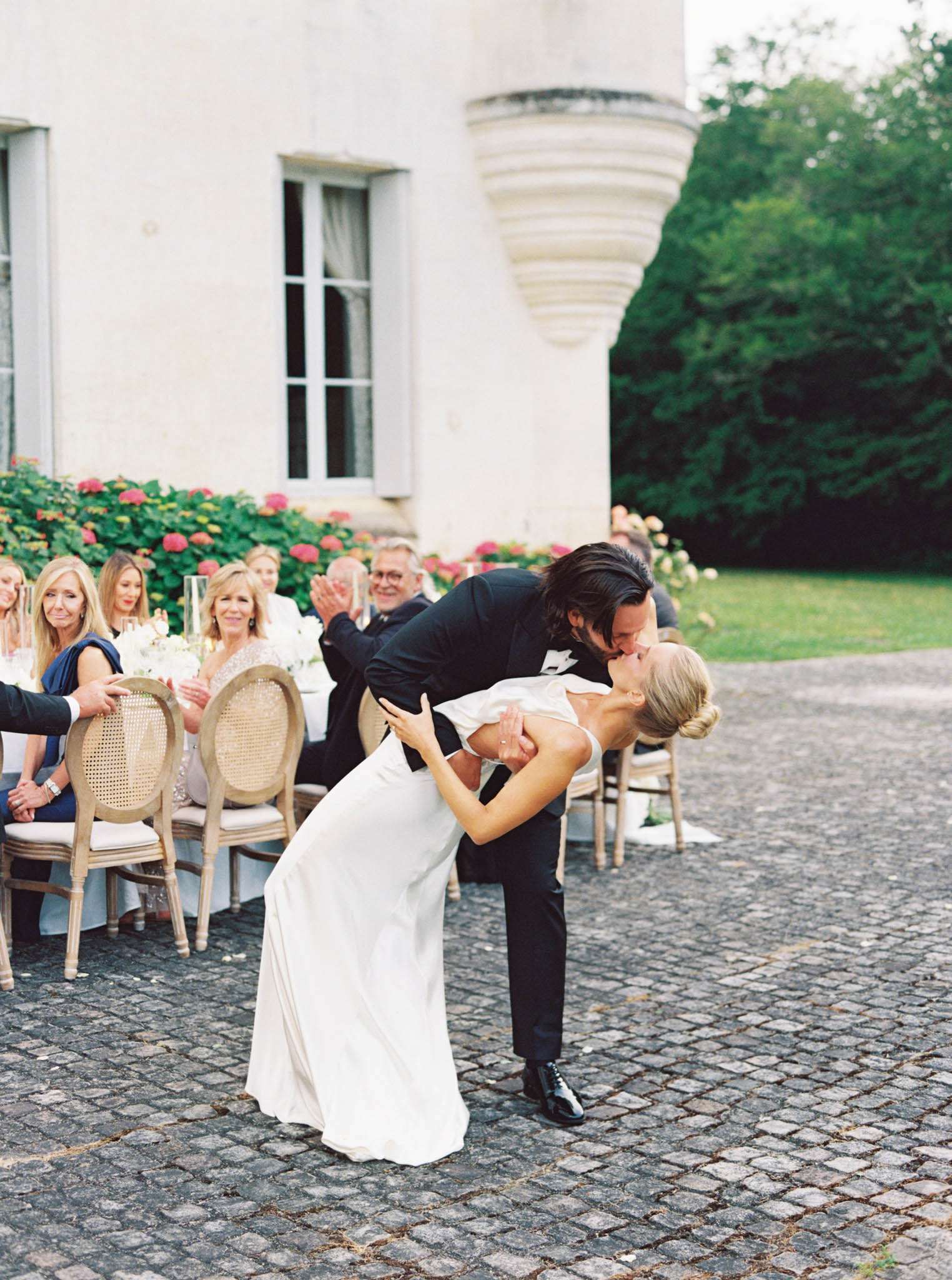 The groom, dressed in a black tuxedo with a bow tie, dips and kisses the bride on a cobblestone courtyard in front of a French château during what appears to be an outdoor reception. The bride wears a sleek, ivory sleeveless gown with a low back and her hair is styled in an updo. Behind them, approximately 10–12 guests are seated at a long banquet table set with white floral arrangements, glassware, and ivory linens, using natural wood cane-back Louis XVI-style chairs. Several guests are visibly reacting with smiles and applause, and pink and white flowering shrubs line the base of the château's stone facade. The shot is a medium wide portrait capturing both the couple's movement and the lively guest reaction in the background.