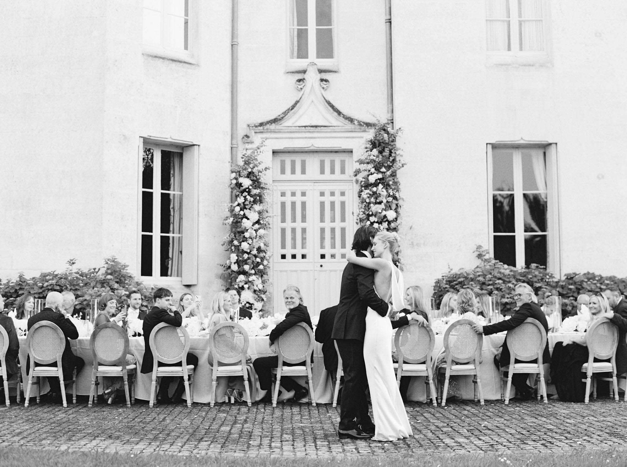 This black-and-white image captures a couple sharing a first dance during an outdoor reception at a French château. The bride wears a sleek, low-back gown while the groom is dressed in a dark suit; they embrace closely in front of the venue's main entrance doors, which are flanked by two tall floral installations of full, lush blooms climbing upward on either side. Behind the couple, a long banquet table seating approximately 25–30 guests stretches across the width of the frame, dressed in light-toned linens with candlestick centerpieces and cane-back Louis XVI-style chairs. The wide shot is composed symmetrically, centering the couple against the château facade, with the seated guests and formal table arrangement framing them on both sides. Potential venue feature image.