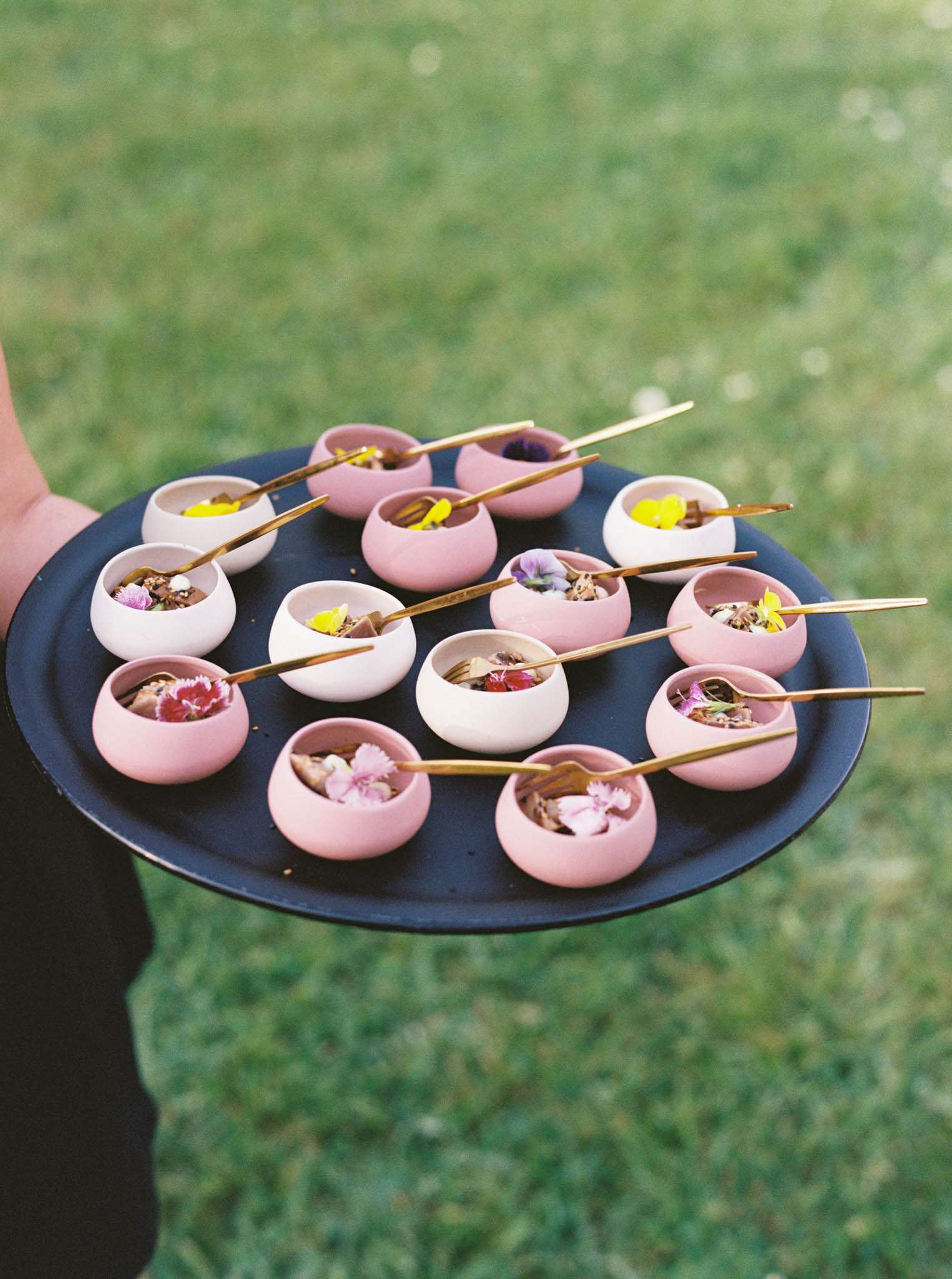 Close-up detail shot of a catering pass during an outdoor cocktail hour, with a server holding a dark navy round tray containing individual amuse-bouche portions served in small rounded ceramic bowls in two colorways — matte white and dusty rose pink. Each bowl contains a small bite garnished with edible flowers in yellow, pink, purple, and red, with gold cocktail picks or mini forks resting across the rims. The tray holds approximately twelve portions arranged in a circular pattern. The styling reflects a modern, design-conscious catering aesthetic with the intentional pairing of the two bowl colors and gold metallic cutlery against the dark tray.