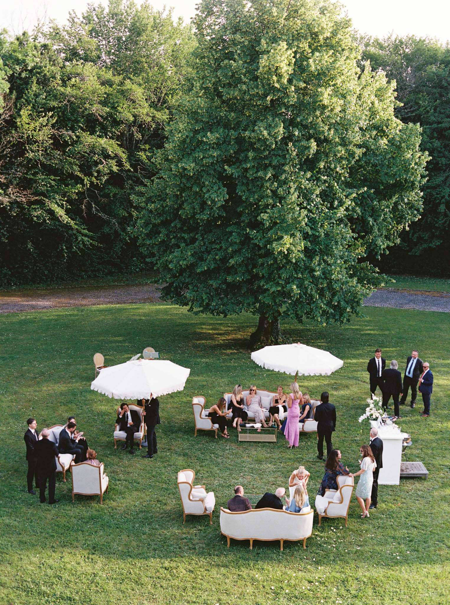 An outdoor cocktail hour is taking place on a manicured lawn, photographed from an elevated wide-angle perspective showing approximately 25–30 guests mingling and seated in lounge areas. The seating arrangement features French Louis-style sofas and armchairs upholstered in ivory fabric with gold-painted wooden frames, grouped around two large white market umbrellas with scalloped edges and a low acrylic coffee table. A white pedestal bar with a white floral arrangement of what appears to be white blooms and greenery is positioned to the right. Guests are dressed in formal attire — men in dark suits and ties, women in cocktail dresses in colors including lavender, blush pink, and black — and are standing in conversation groups or seated across the lounge clusters. The styling theme is classic French with a refined, garden-party aesthetic combining the ornate vintage furniture with the white umbrella canopies.