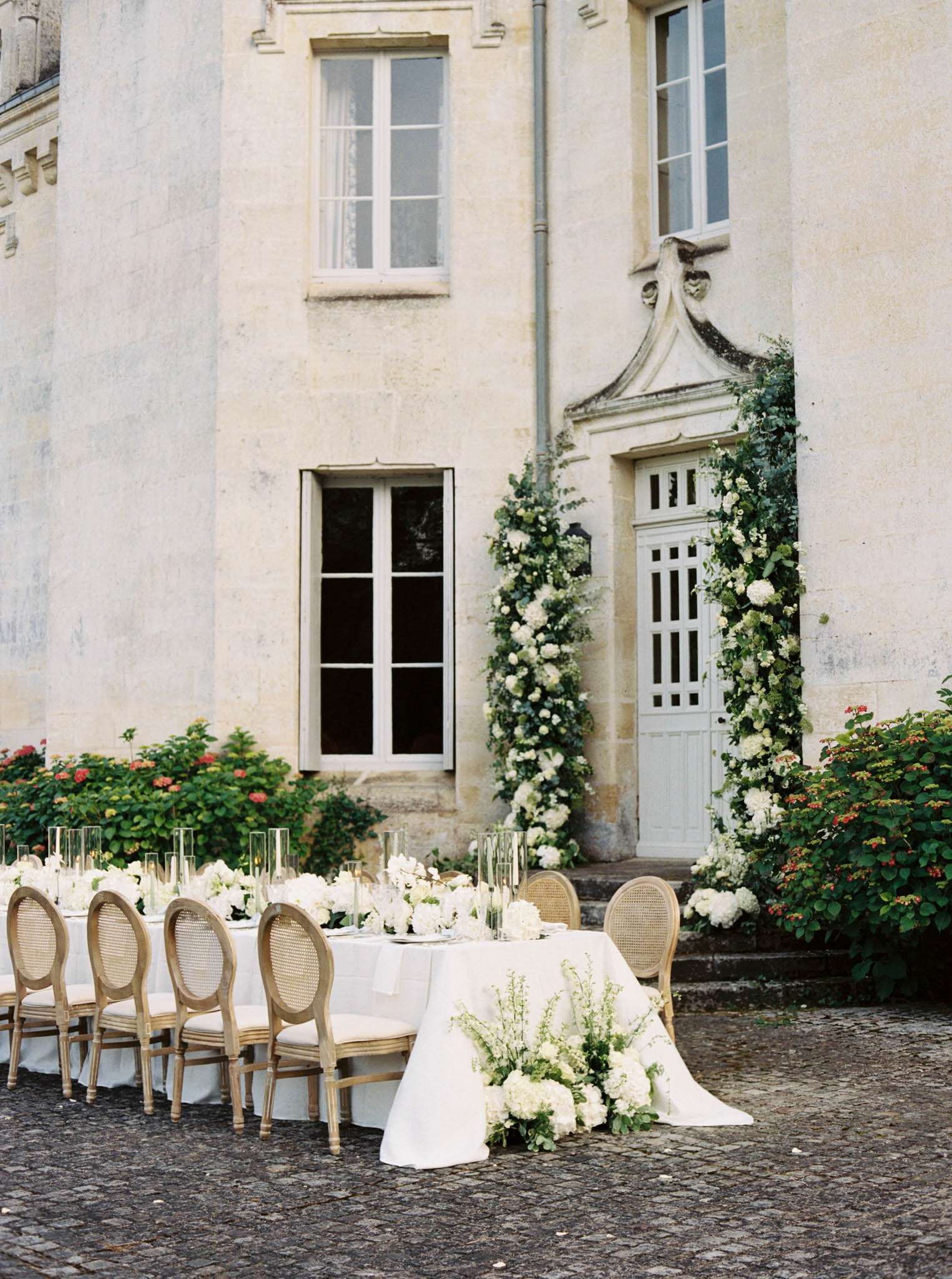 Long reception table with white hydrangea runner and floral arch framing chateau doorway