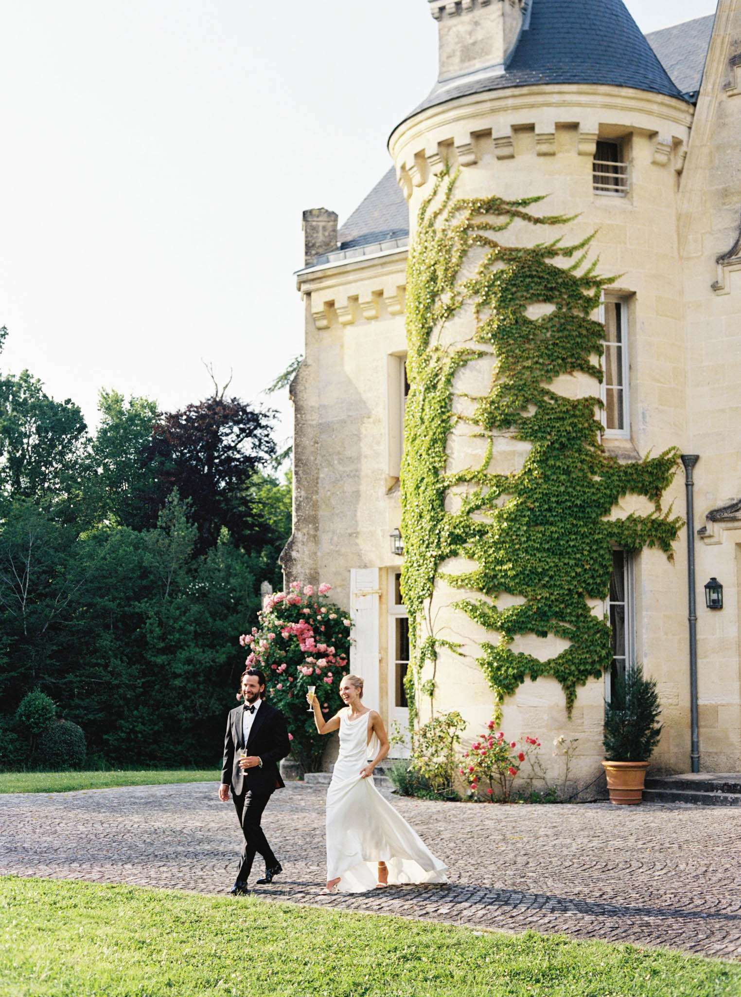 Bride and groom walking with champagne glasses on cobblestone grounds of ivy-covered chateau tower