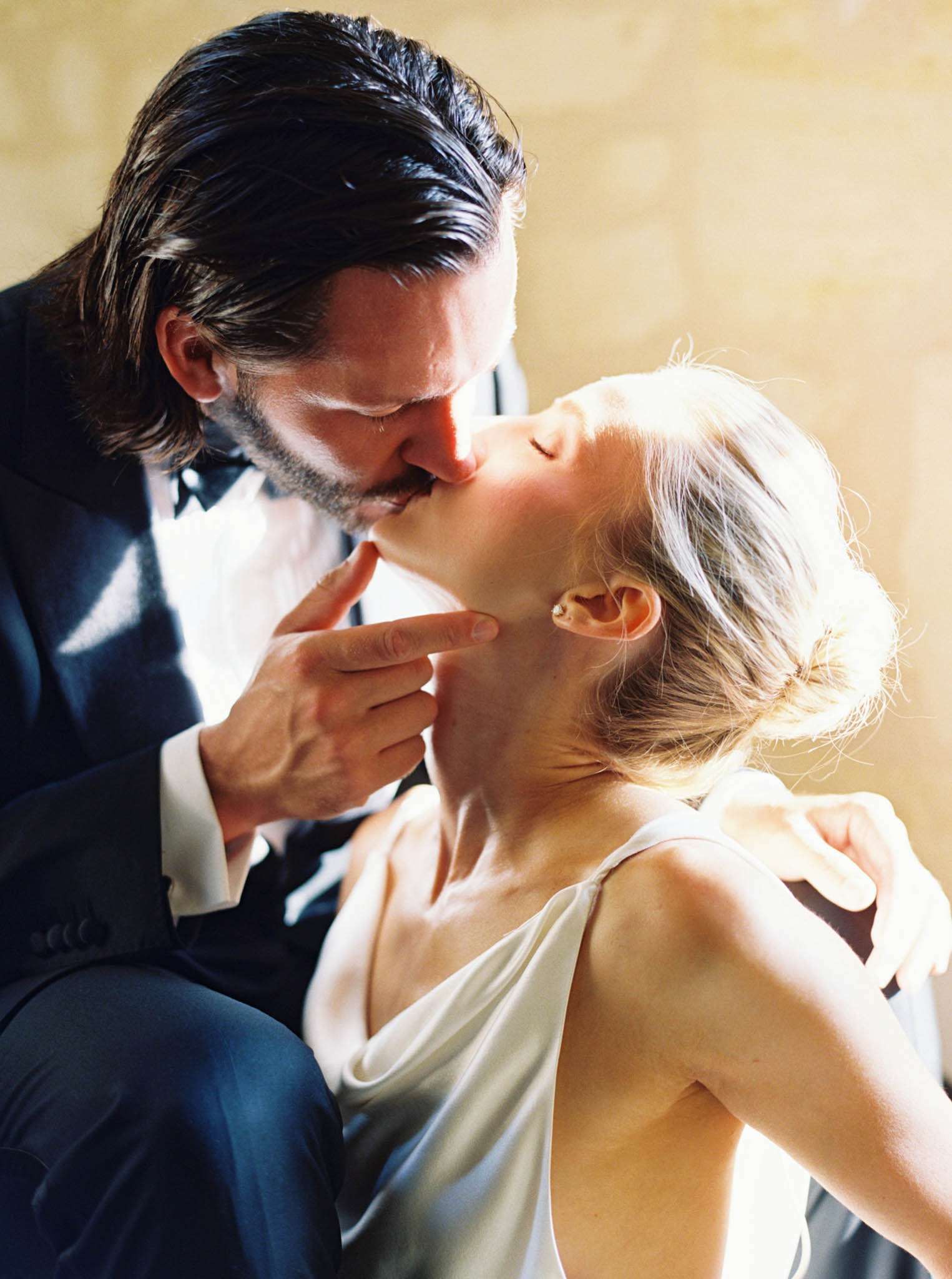 Close-up of groom in navy tuxedo kissing bride in ivory cowl-neck gown with warm golden light
