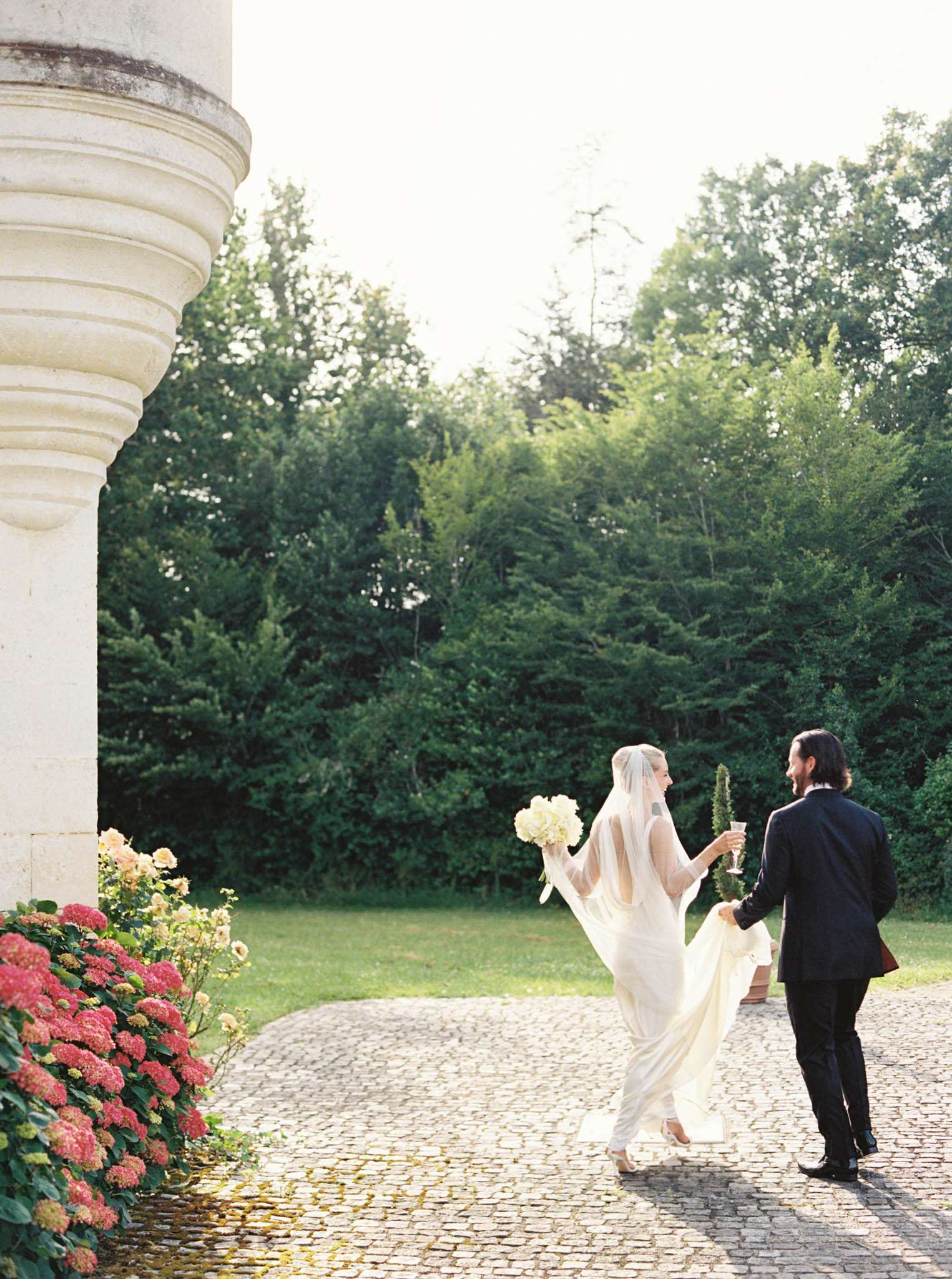 Bride and groom walking along cobblestone chateau path from behind, bride with cathedral veil and cream bouquet