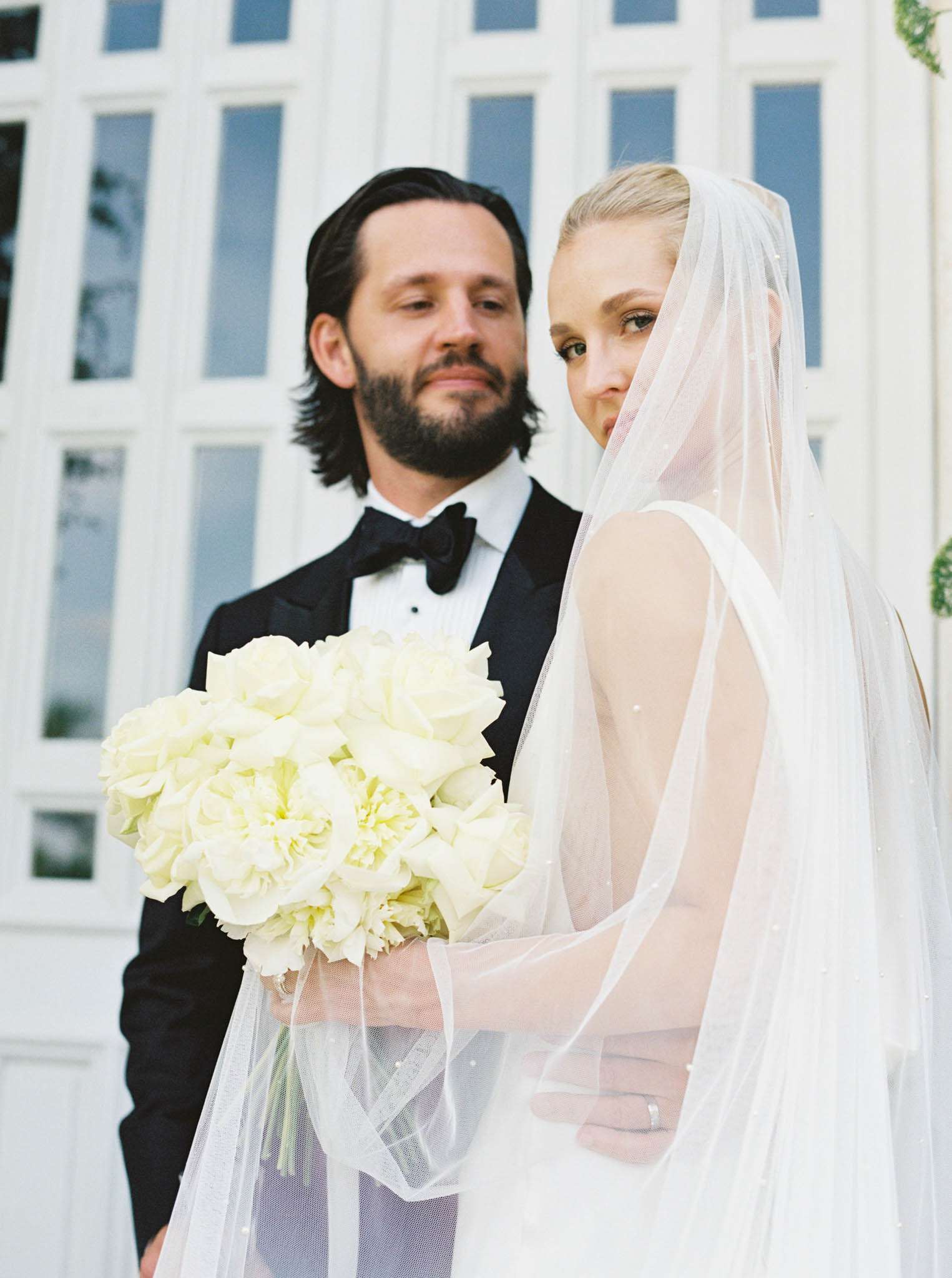 Bride in backless gown with pearl-embellished veil holding white rose bouquet beside groom in black tuxedo