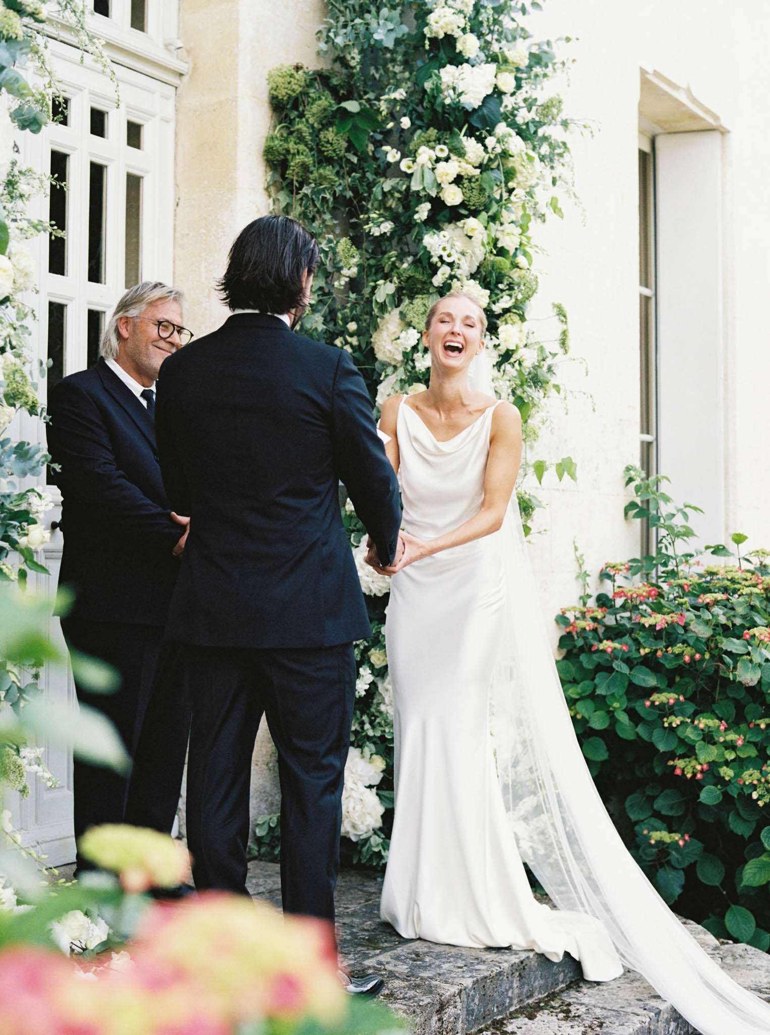 An outdoor ceremony moment captured at the entrance of a French château, where the bride and groom stand face to face holding hands while an officiant looks on with a smile. The bride wears a sleek ivory cowl-neck slip gown with a long tulle veil, and is laughing openly with her eyes closed. The groom and officiant are both dressed in dark navy suits. Behind them, a large floral installation climbs the château facade, composed of white hydrangeas, white ranunculus, green viburnum, and trailing greenery in a lush, organic arrangement. The shot is taken from a medium distance with foreground foliage softly blurring into the frame, giving depth to the composition. The overall styling is modern and clean, with a green-and-white floral palette against the pale stone building.