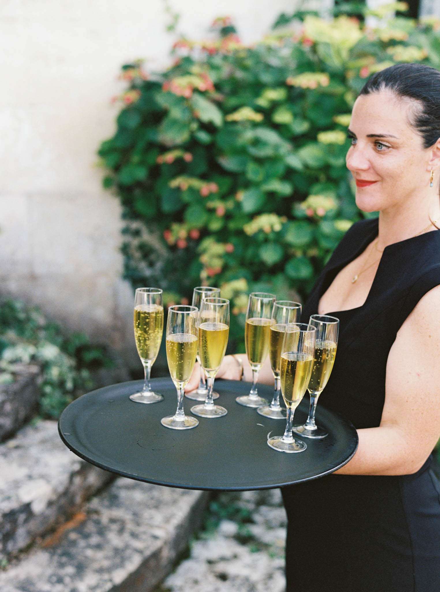 Server carrying tray of champagne flutes against stone wall with climbing foliage at chateau cocktail hour