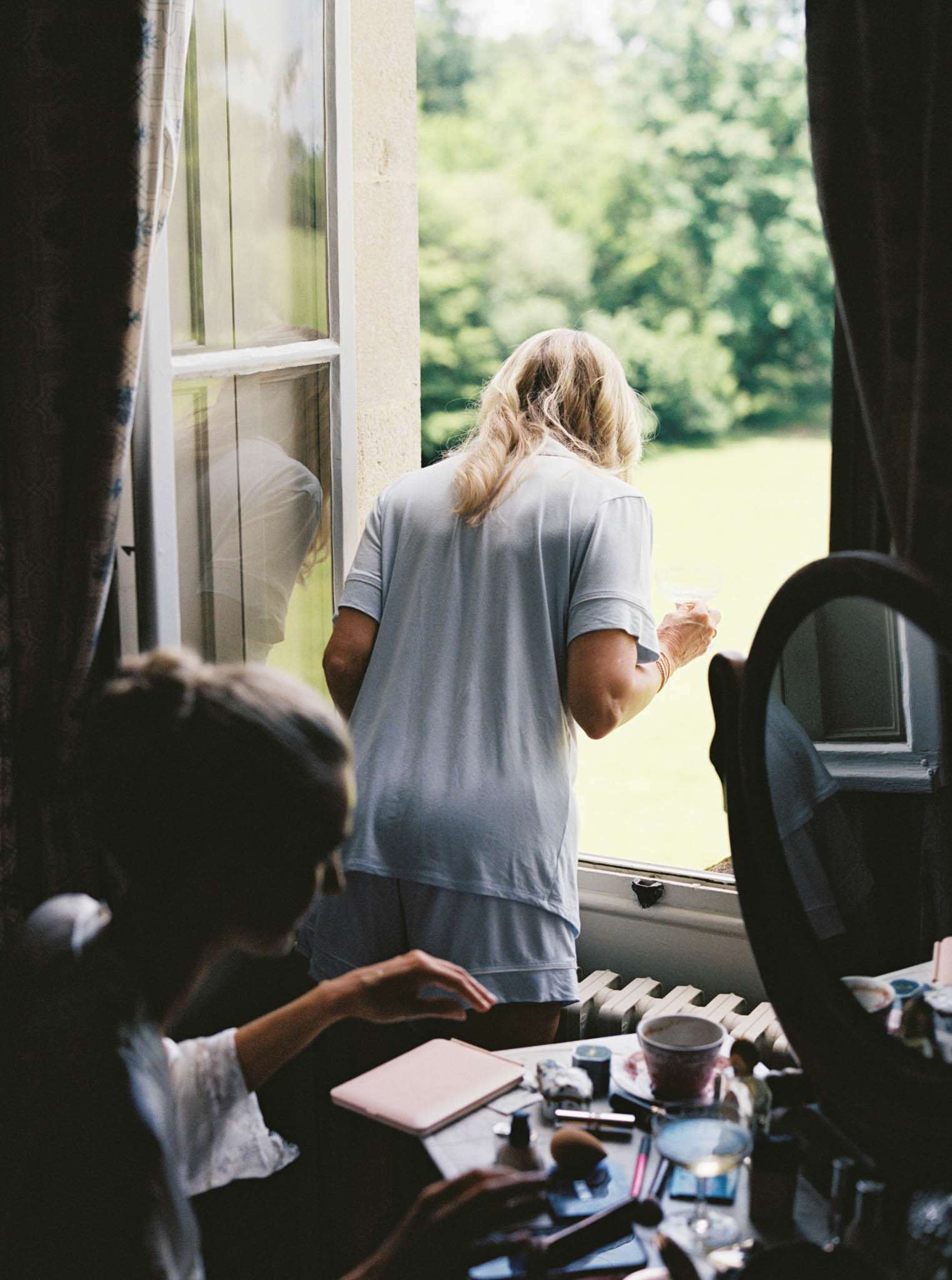 A getting-ready scene set in an indoor room, likely within a château or manor house, identifiable by the tall French-style casement windows with white frames and heavy dark curtains with a floral lining. One woman with long blonde hair, wearing a pale blue short-sleeve pajama set, stands at the open window holding a small glass and looking out toward a manicured lawn. A second woman, seen from behind and slightly out of focus in the foreground, sits at a dressing table covered in makeup products including brushes, compacts, a pink notebook, and various cosmetic items, with a round black-framed mirror to the right. The composition is a medium candid shot taken from behind both subjects, giving an intimate, unposed feel to the pre-ceremony preparations.
