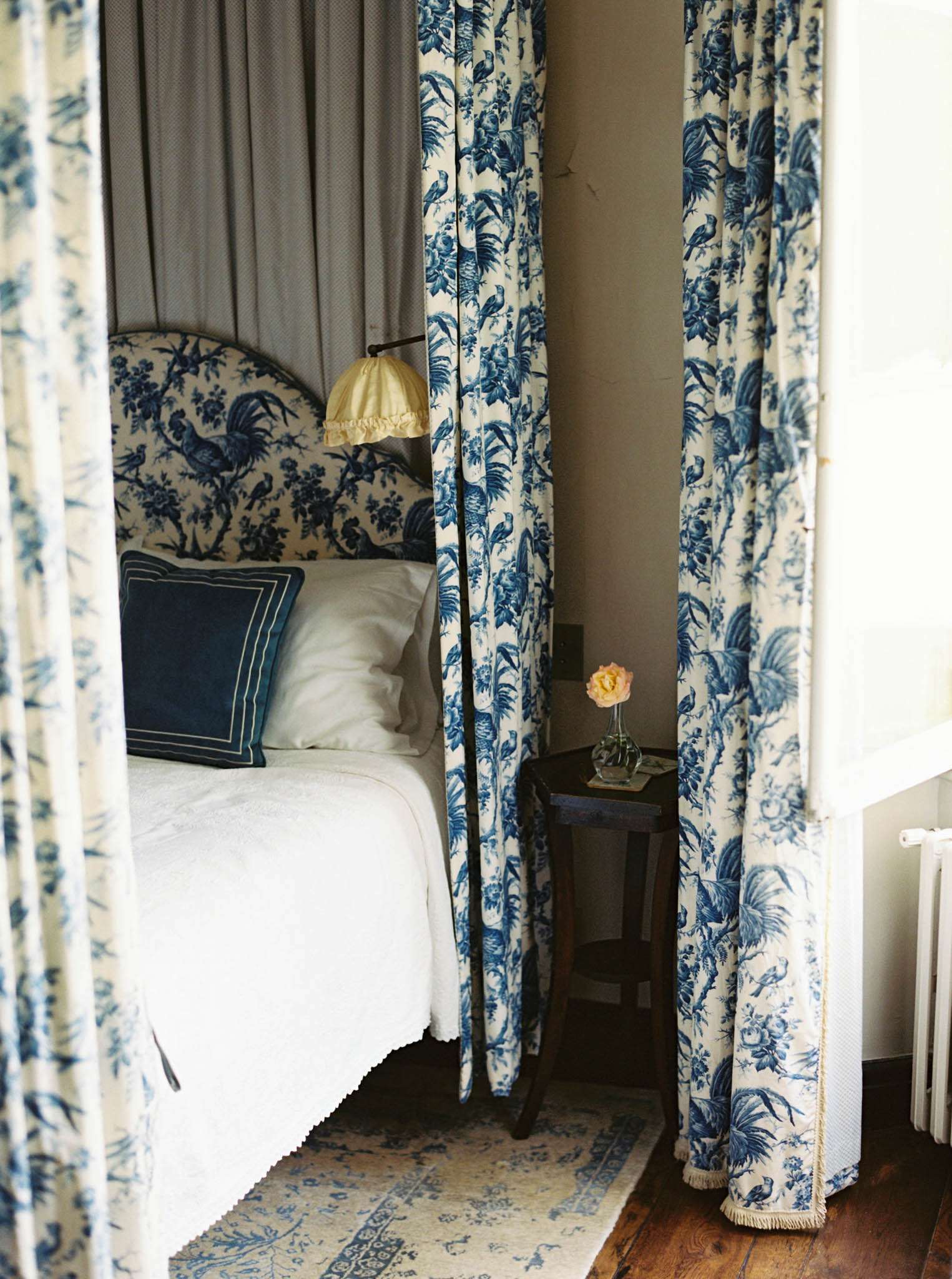 An interior detail shot of a bedroom, likely within a château or historic venue used for a wedding weekend. The room features blue-and-white toile de Jouy fabric used consistently across the curtains and upholstered headboard, depicting birds and roosters on a cream background. The bed is dressed in white linens with navy blue bordered cushions, and a fringed blue-and-cream Oriental rug is visible on the dark hardwood floor. A small dark wood bedside table holds a single peach rose in a glass bud vase, accompanied by a yellow-shaded wall-mounted lamp. The overall styling is classic French country in a navy and cream palette. Potential venue feature image.