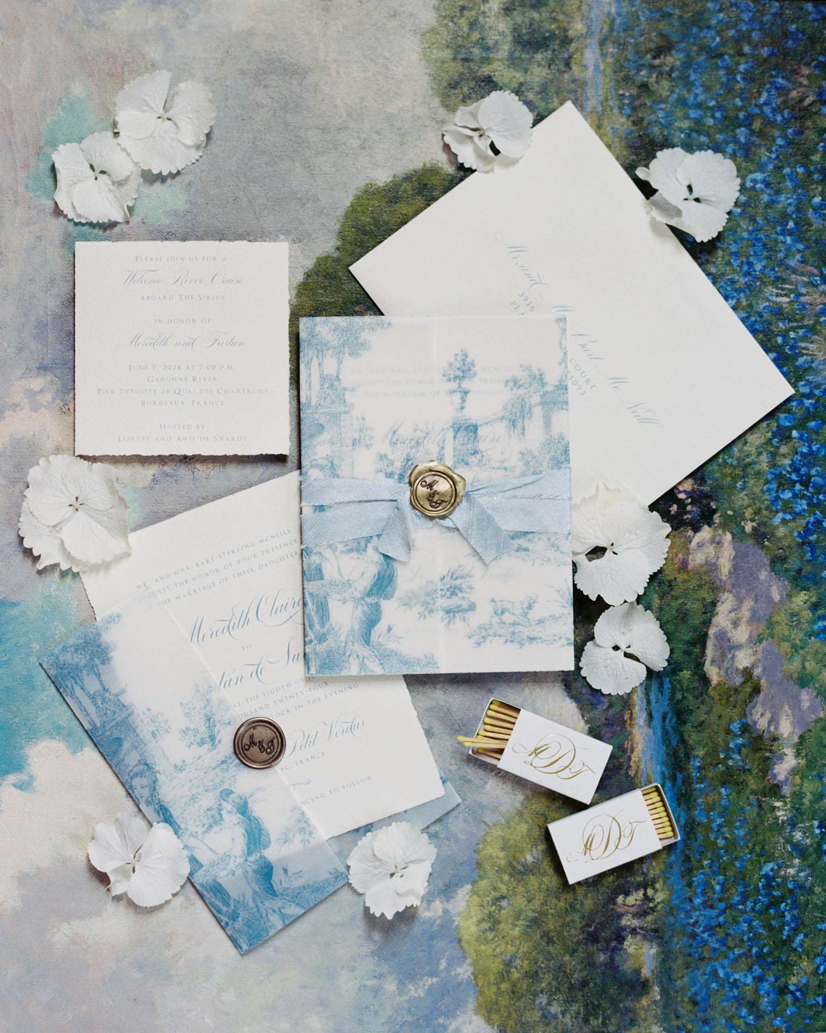 A flat lay detail shot of a wedding stationery suite displayed on an impressionist-style painted canvas backdrop featuring soft blue, green, and white tones. The suite includes a main invitation card with blue toile de Jouy printed paper wrap tied with a pale blue silk ribbon and sealed with a gold wax seal stamped with monogram initials, a formal ceremony invitation card with calligraphy script, a separate welcome river cruise card, an envelope with calligraphy addressing, and two custom monogrammed matchboxes with gold-tipped matches. White hydrangea florets are scattered loosely around the stationery pieces as styling accents. The overall color palette is powder blue, ivory, and gold, with a classic French toile print as the central design motif.