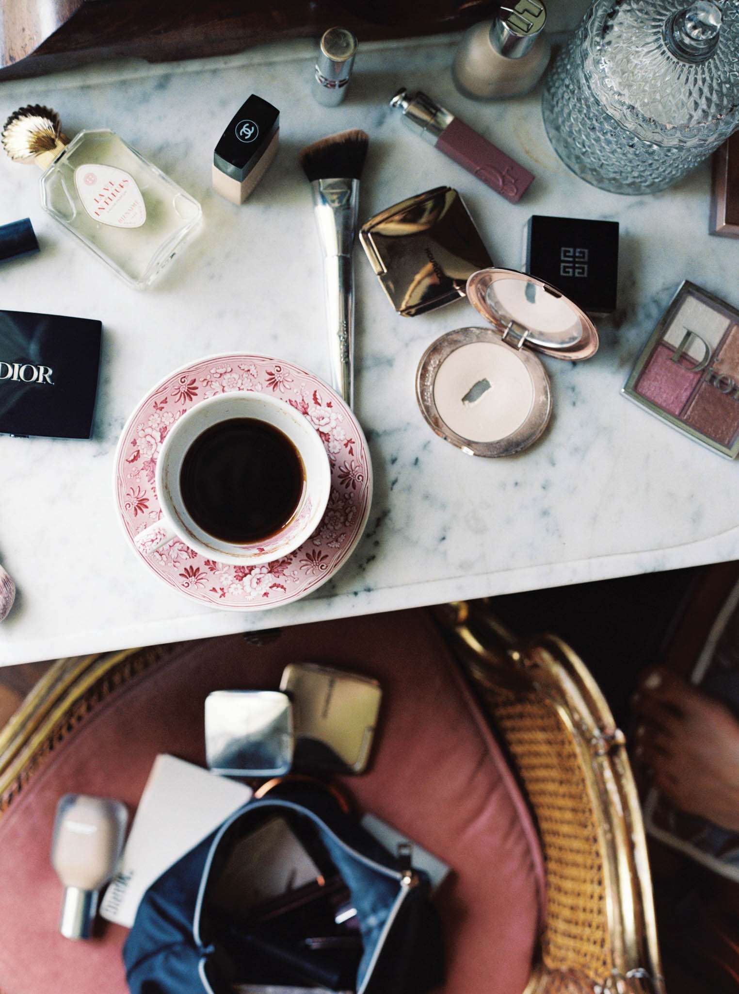 A flat-lay detail shot taken from above during a bridal getting-ready scene, showing a white marble-topped vanity table scattered with luxury makeup products including Dior, Givenchy, and Chanel compacts and palettes, a gold compact mirror, makeup brushes, a perfume bottle, and a mauve lipstick. A pink and white floral-patterned china cup and saucer holding black coffee sits at the center of the arrangement. Below the marble surface, a dusty pink upholstered chair holds an open navy blue makeup bag with additional products, alongside a woven rattan and gold handbag. The styling reflects a classic, French-influenced aesthetic with high-end beauty brands and vintage-style chinaware as intentional props.