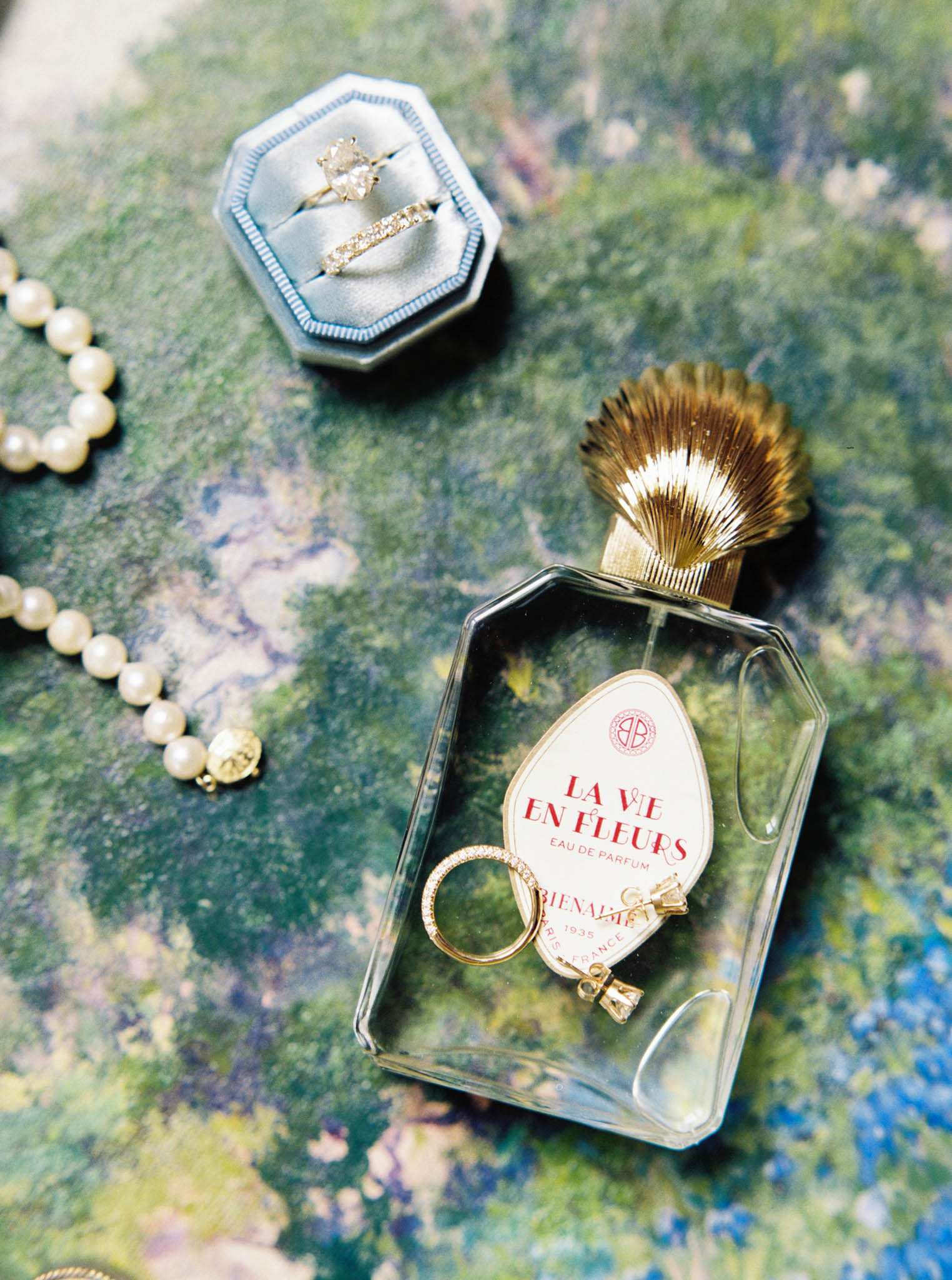 A close-up flat lay of bridal accessories arranged on a green and blue marbled surface. A dusty blue octagonal velvet ring box holds two rings — a solitaire engagement ring with a pear-shaped diamond in a gold setting and a diamond eternity band. Nearby, a pearl necklace with a gold clasp is partially visible along the left edge. A glass perfume bottle labeled 'La Vie en Fleurs Eau de Parfum' by Bienaimé, Paris, France (1935), with a gold scallop shell cap, sits in the center foreground, and inside the glass tray of the bottle's base rest two additional gold rings — one a diamond pavé band and one a small bow-shaped piece. The styling has a French vintage aesthetic with a warm gold and blue color palette.