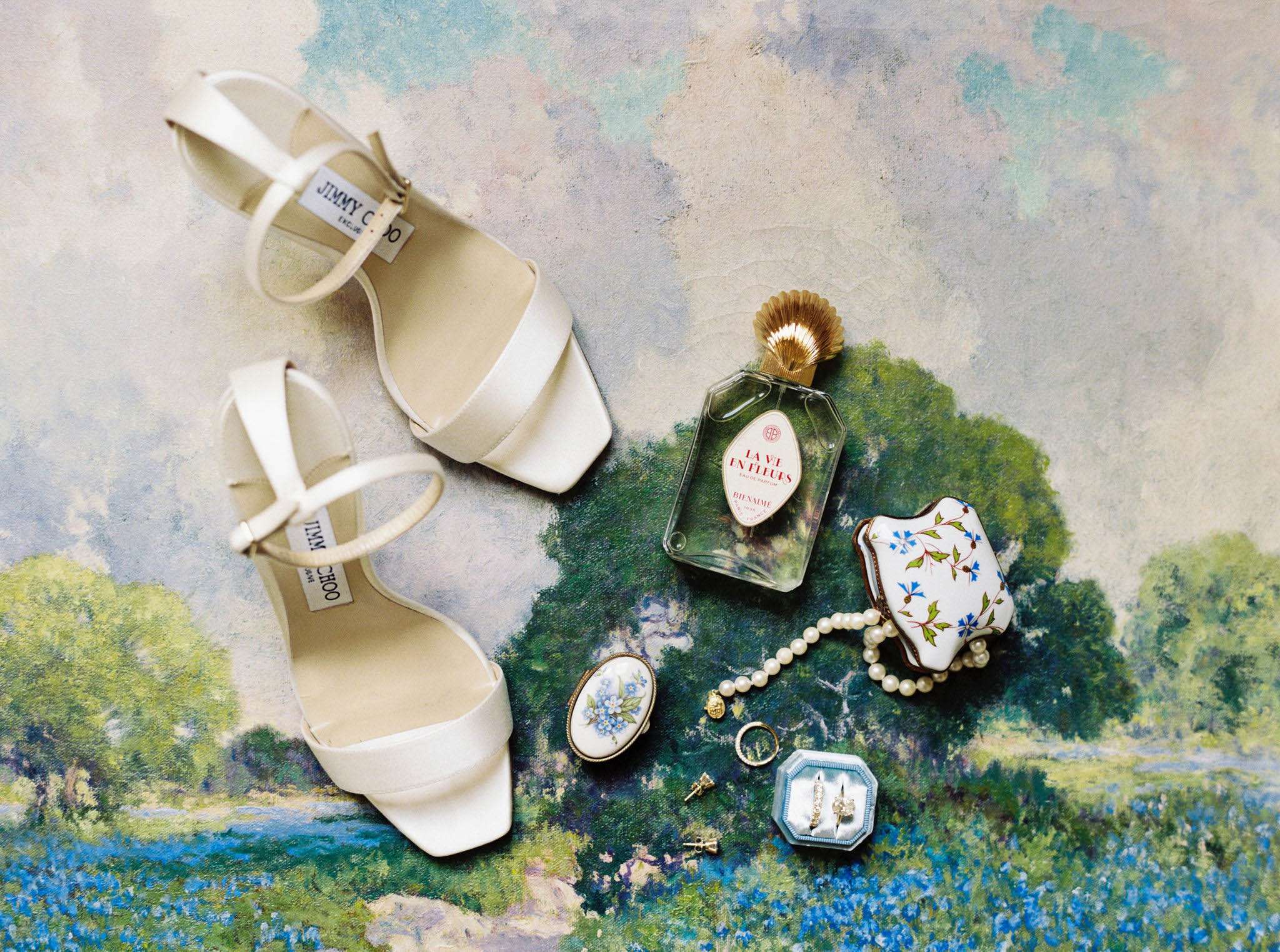 Bridal flat lay with ivory Jimmy Choo sandals, pearl bracelet, blue ring box, and vintage perfume on painted background