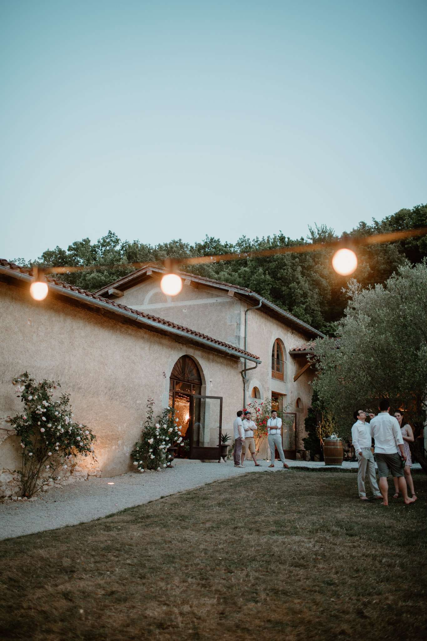 Guests mingling at dusk on lawn before Provencal stone building with globe string lights and climbing roses