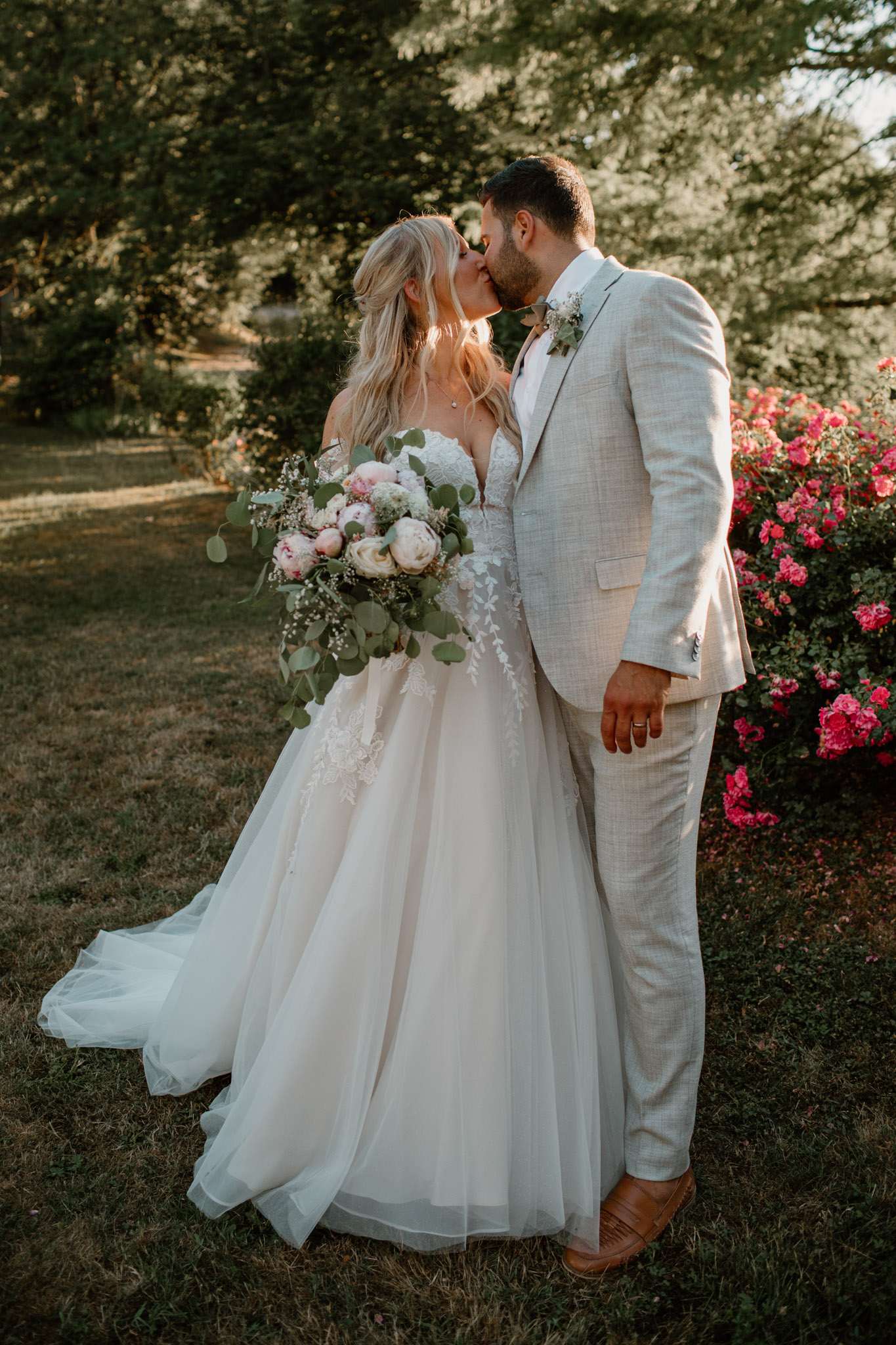 Couple kissing at golden hour, bride with blush peony bouquet in lace applique gown, groom in sand suit