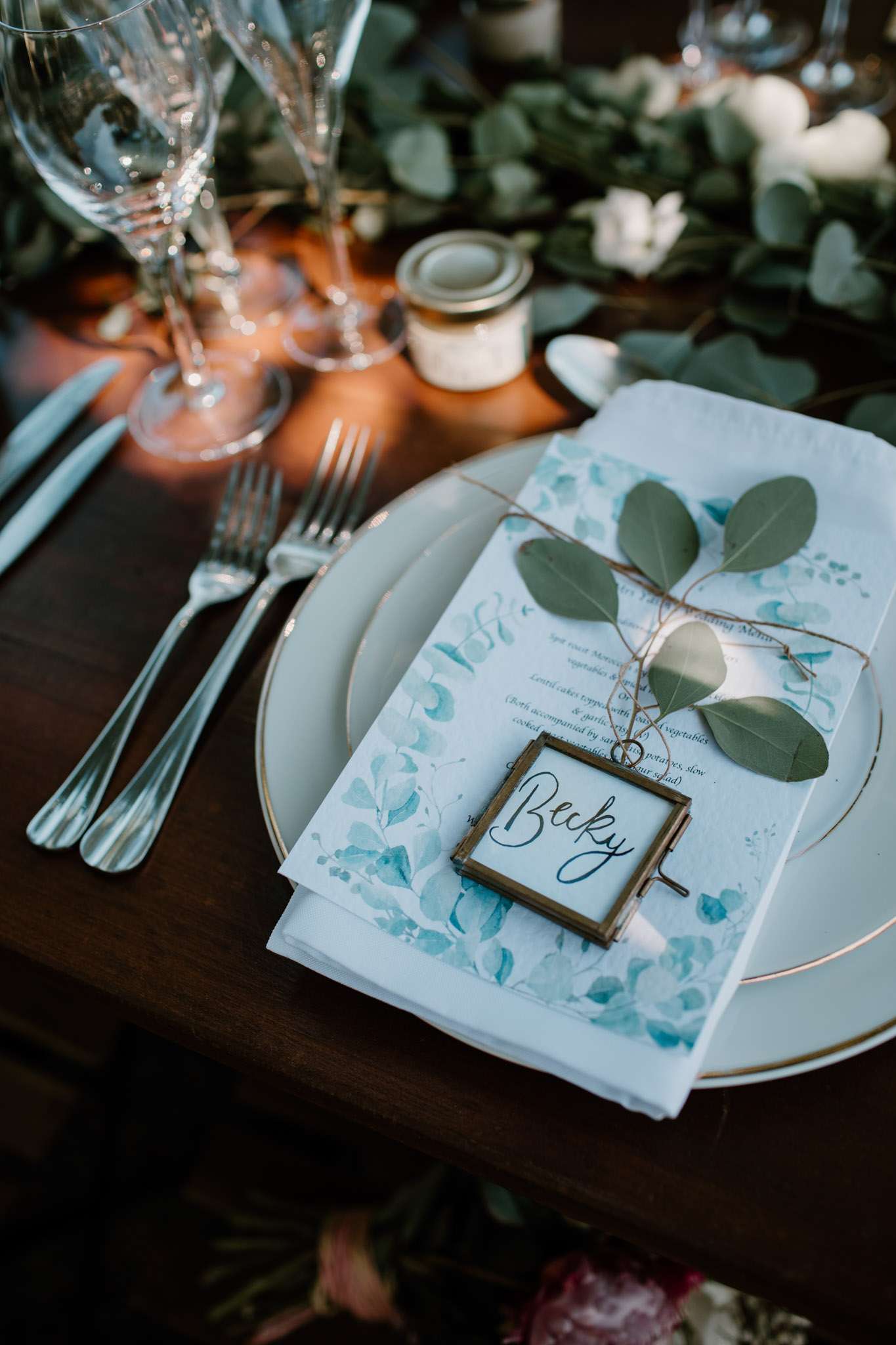 Place setting with eucalyptus menu card, calligraphy name card, gold-rim plate, and green garland centerpiece