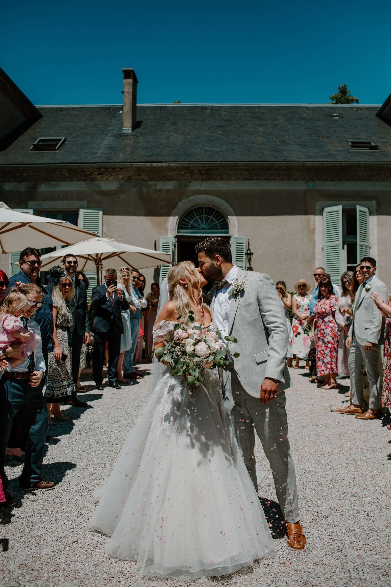 Bride and groom kiss during confetti exit on gravel courtyard before French manor with guests cheering