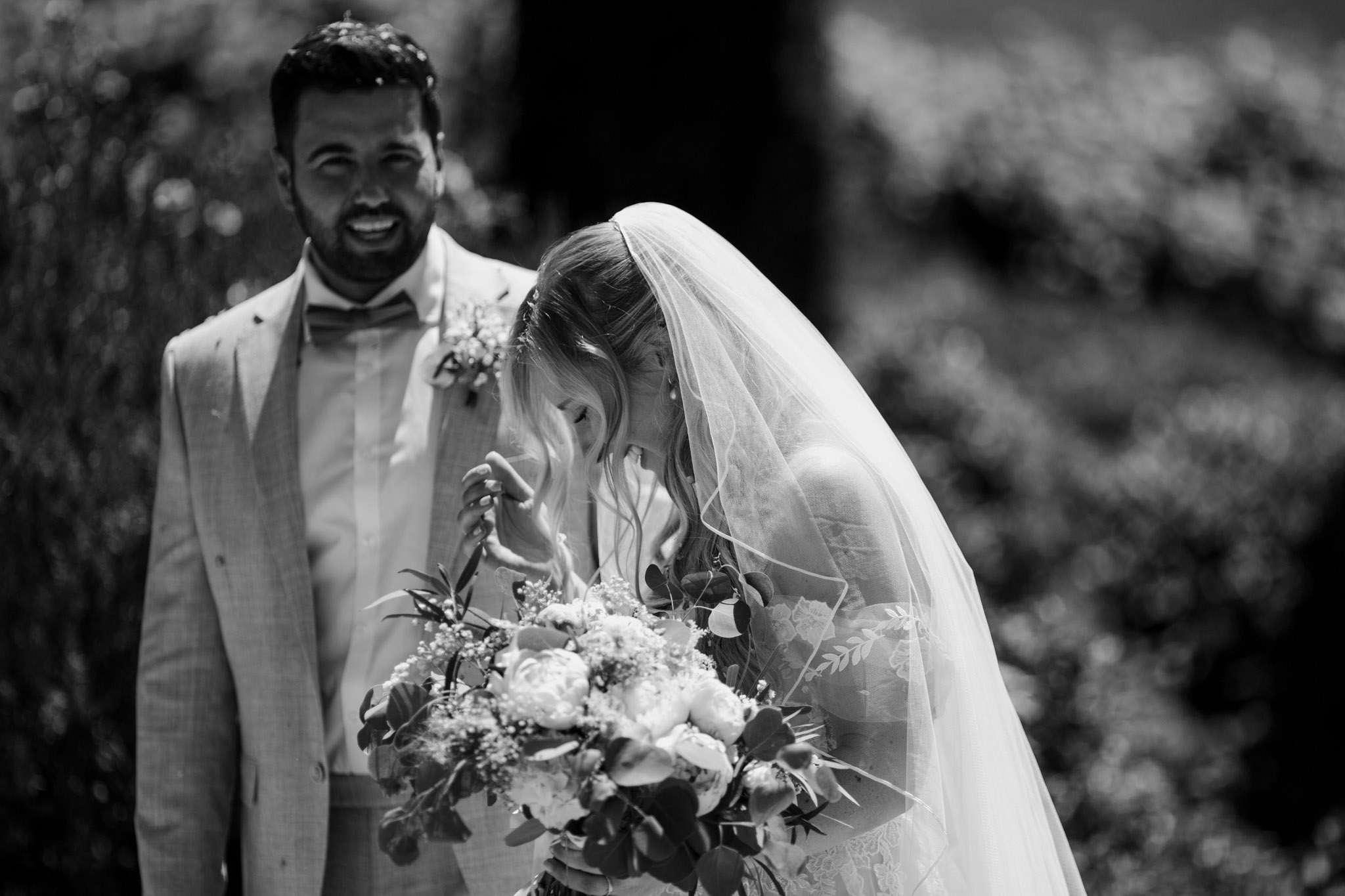 This black-and-white portrait captures what appears to be a first look or an emotional moment between the bride and groom outdoors, with soft dappled light and a blurred leafy background. The bride is bowing her head and wiping away tears with one hand while holding a large bouquet of peonies, small filler flowers, and trailing foliage with the other; she wears a lace-detailed dress and a flowing veil. The groom stands just behind her, laughing broadly with evident joy, dressed in a light-colored suit with a bow tie and a small floral boutonnière on his lapel. The image is a close-up two-shot with strong contrast between the bright highlights on the couple and the darker background foliage.