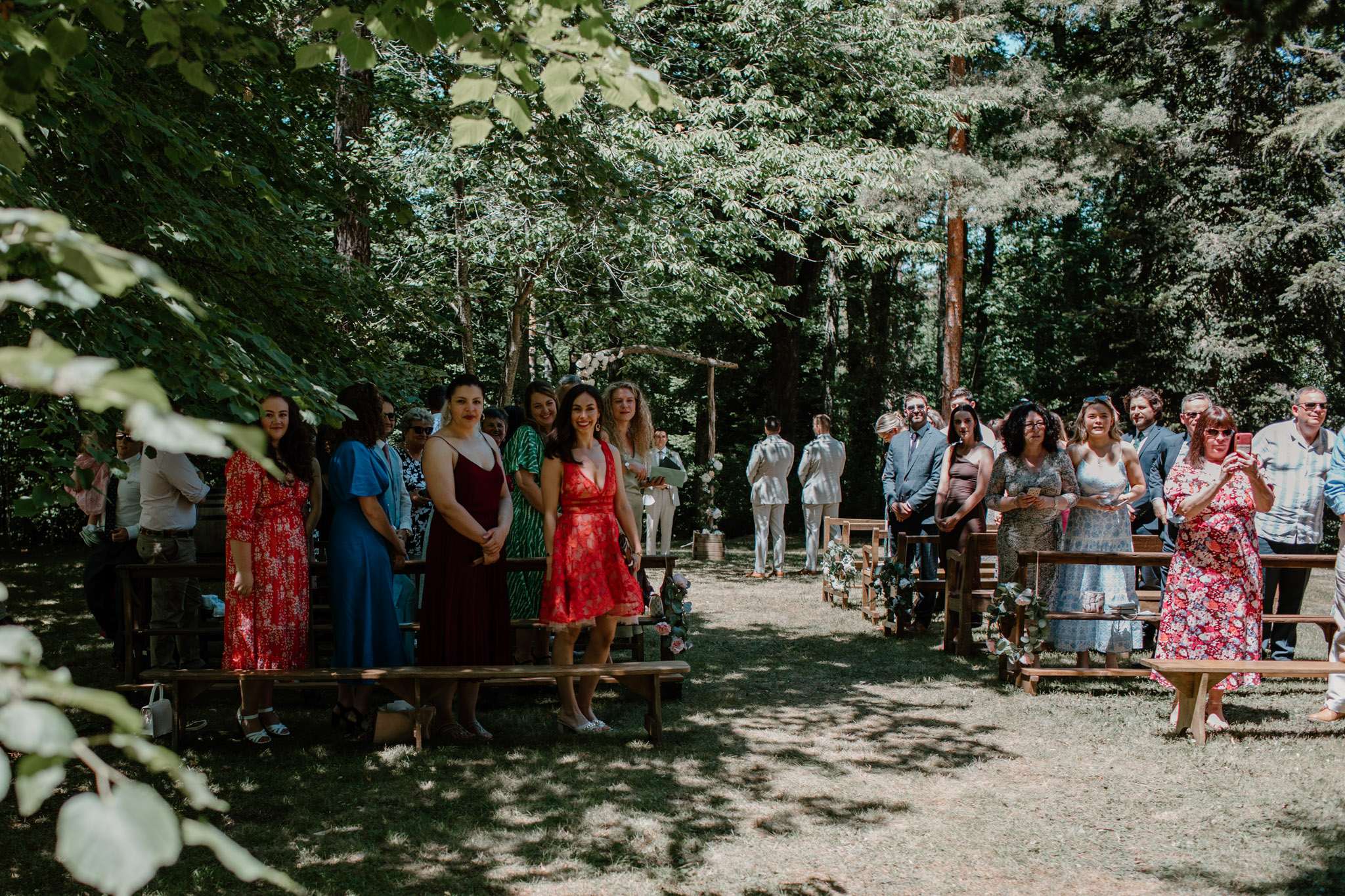 An outdoor woodland wedding ceremony in progress, with approximately 30–40 guests standing on either side of a central aisle lined with wooden benches. The aisle is decorated with small floral arrangements featuring blush pink roses and trailing greenery attached to the bench ends. At the far end of the aisle, the groom stands in a white or ivory suit beneath a rustic wooden arch decorated with white florals, facing away from the camera. Guests are dressed in a variety of colorful summer attire including a red lace mini dress, a burgundy midi dress, a teal blue dress, and floral prints, giving the event a relaxed, boho-casual feel. The ceremony setup uses natural wooden elements — log posts and simple timber benches — consistent with a rustic, nature-focused styling theme. The shot is a wide-angle view taken from the back of the aisle looking toward the altar, captured in bright midday light filtering through the tree canopy.