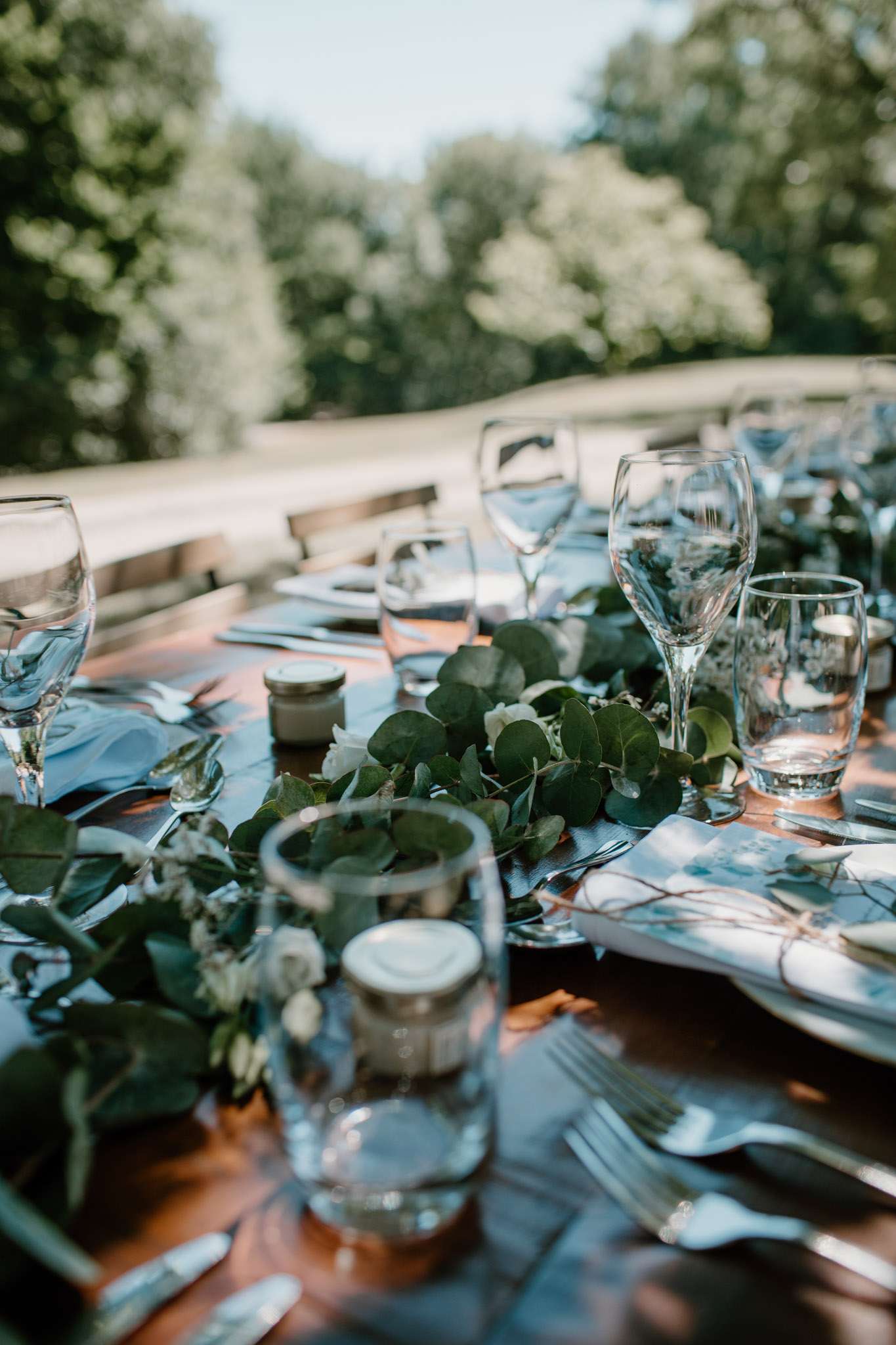 A close-up detail shot of an outdoor wedding reception table set for a meal in natural daylight. The wooden farm-style table is dressed with a lush eucalyptus and white rose garland running down the center, accompanied by small glass tealight holders and individual honey jar favors. Place settings include dusty blue folded napkins, silver cutlery, clear wine and water glasses, and printed menus or place cards tied with twine. The decor palette combines warm wood tones with dusty blue and greenery, reflecting a relaxed garden or rustic-natural styling.