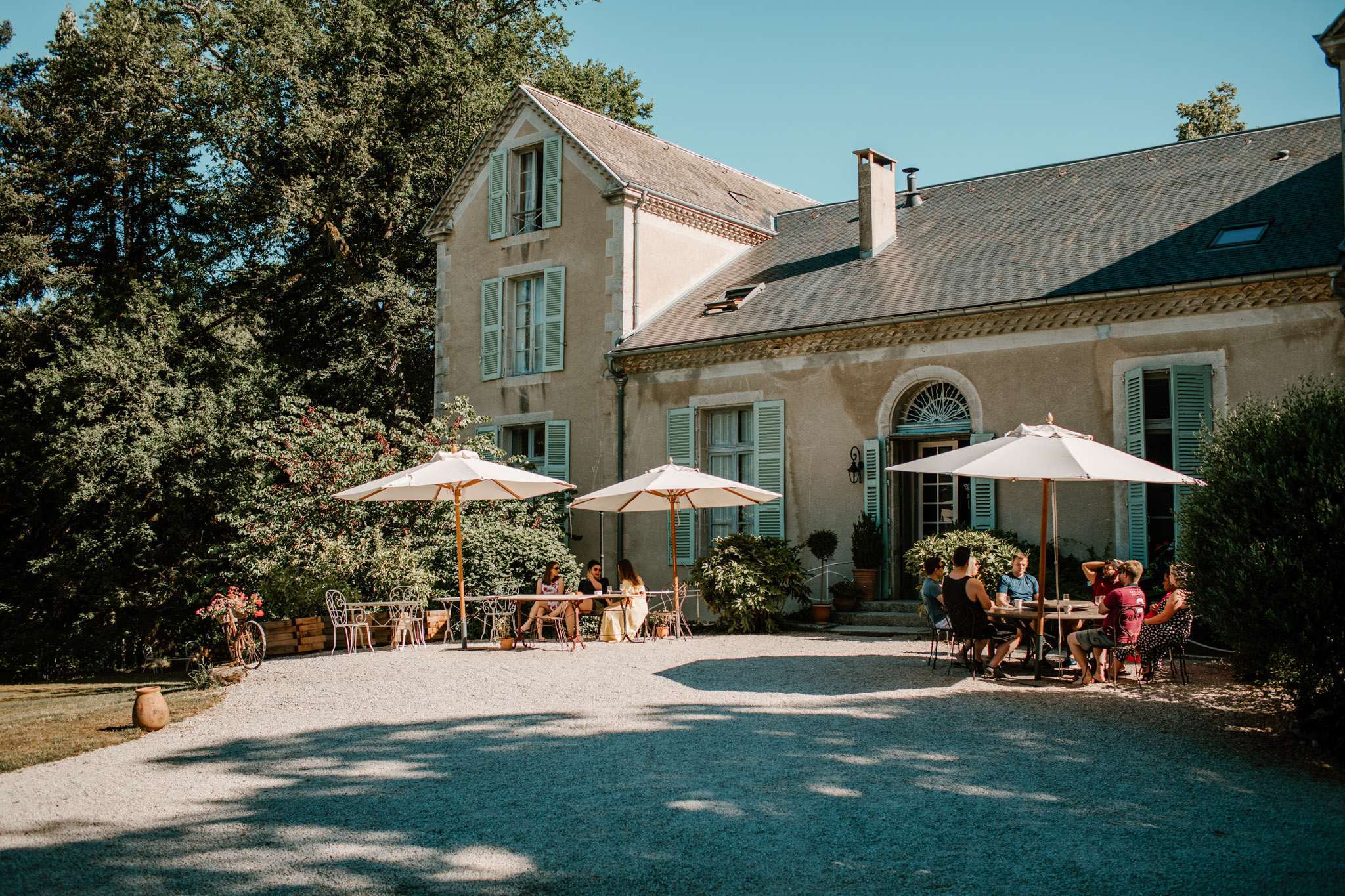 Guests seated under cream umbrellas on gravel courtyard of French manor with sage-green shutters