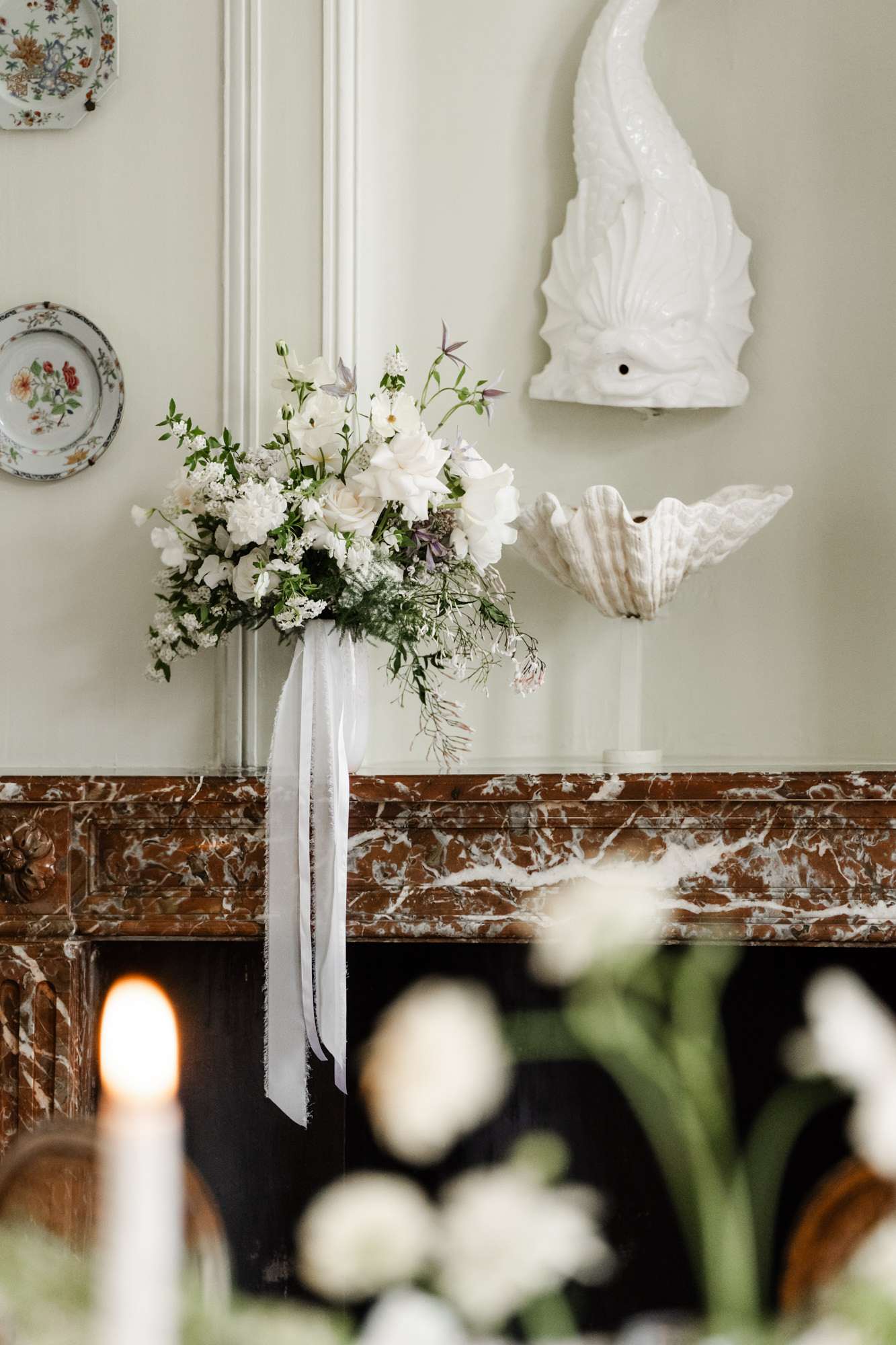 White bridal bouquet with ivory roses, ranunculus, and burgundy accents on marble fireplace mantel with cream molding behind