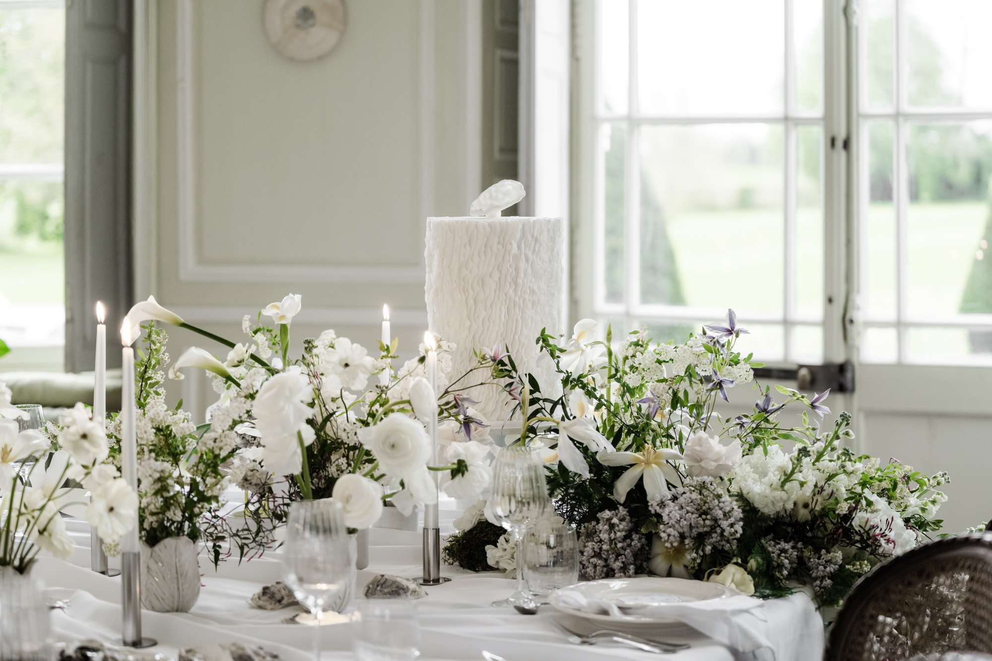 Indoor reception table with white wedding cake flanked by anemone and ranunculus arrangements and taper candles