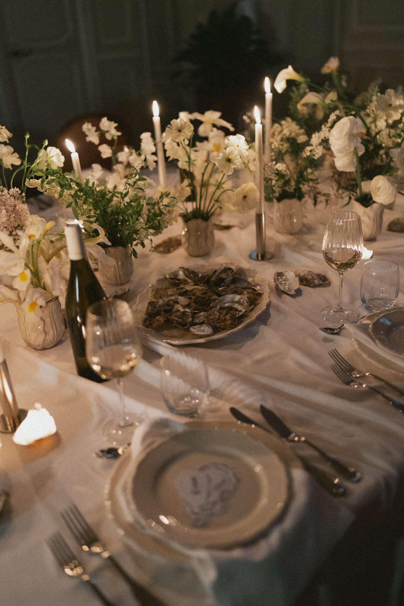 Evening reception table with white ceramic flower vessels, taper candles, oyster plate, and wine bottles