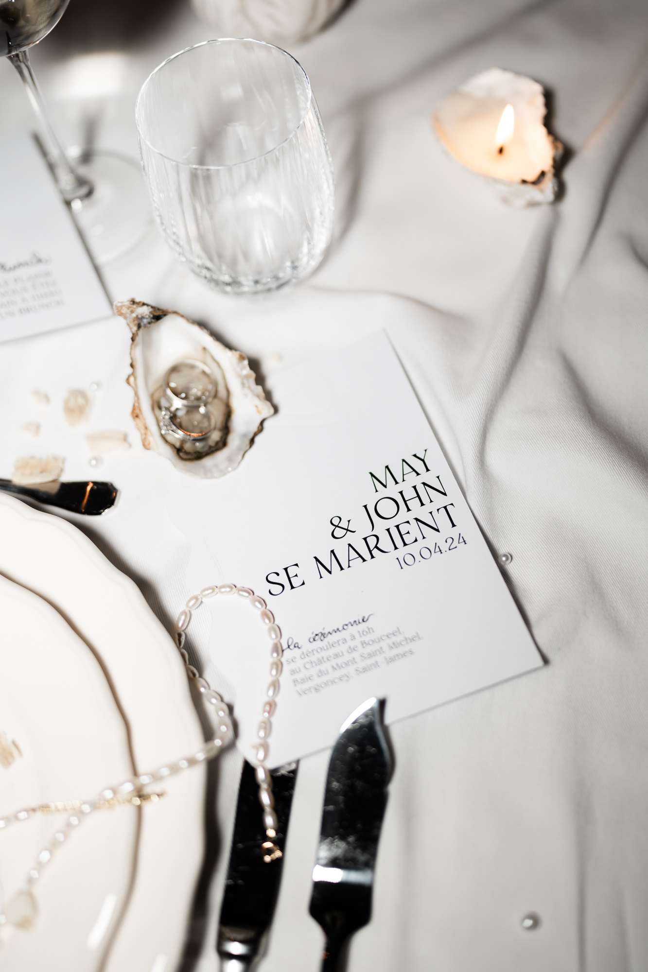 Flat lay of wedding place setting with white linen napkin, black flatware, printed menu card, and rings in oyster shell