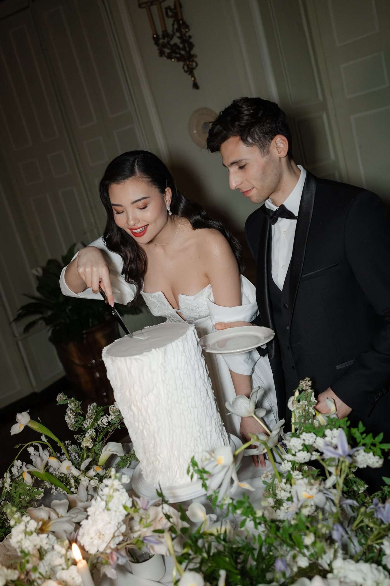 Bride and groom cutting white textured wedding cake surrounded by white daisy and rose arrangements in a minimalist room